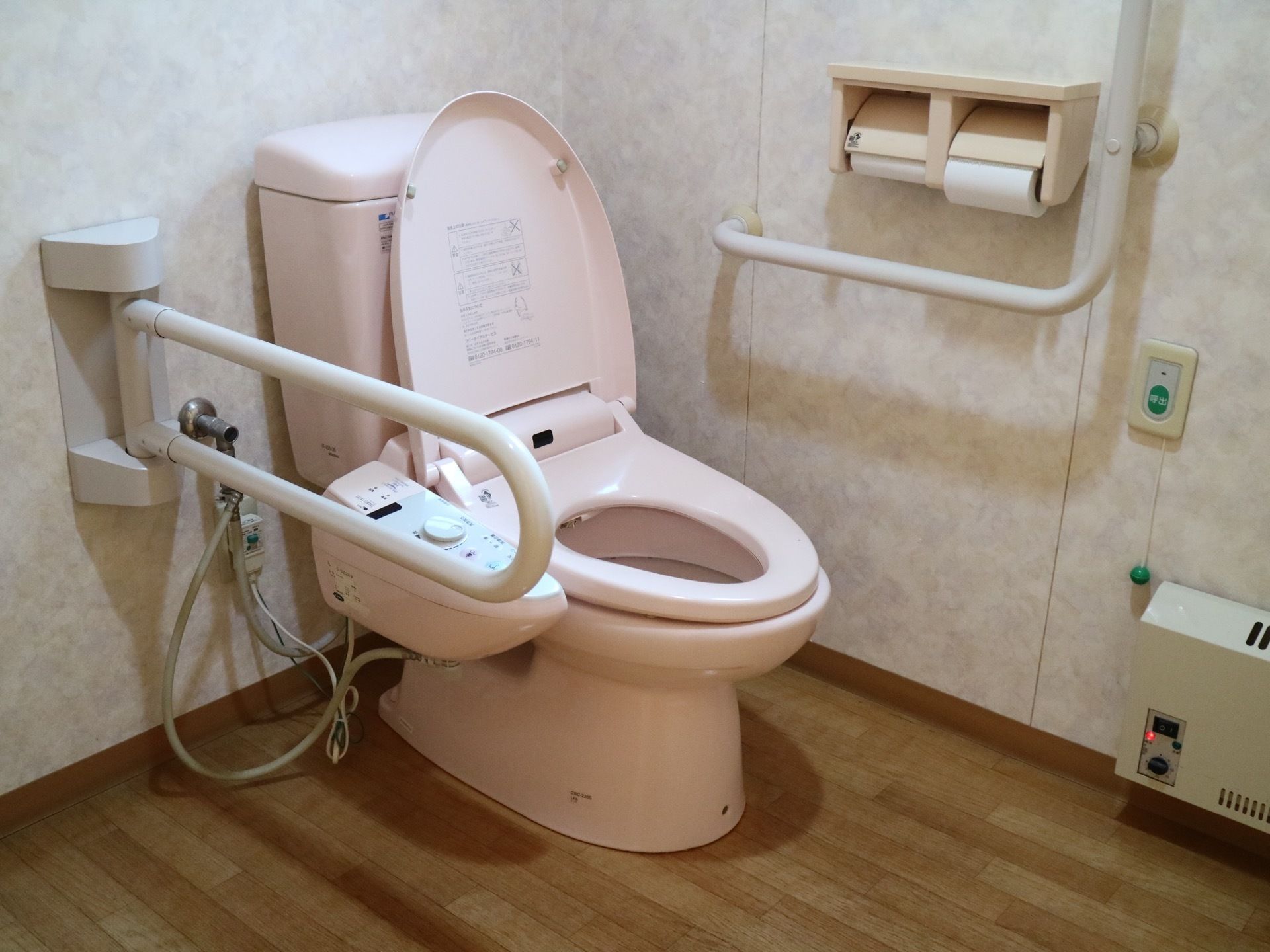 中央に薄いピンク色のトイレがあり、温水洗浄便座が取り付けられている。便座の手前に可動式の手すりがある。
