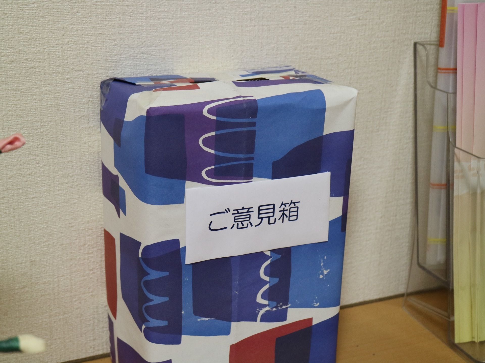 木製の棚の上にご意見箱が置かれている。空き箱を利用しており、青い模様の入った紙で包まれている。右側にラックがある。