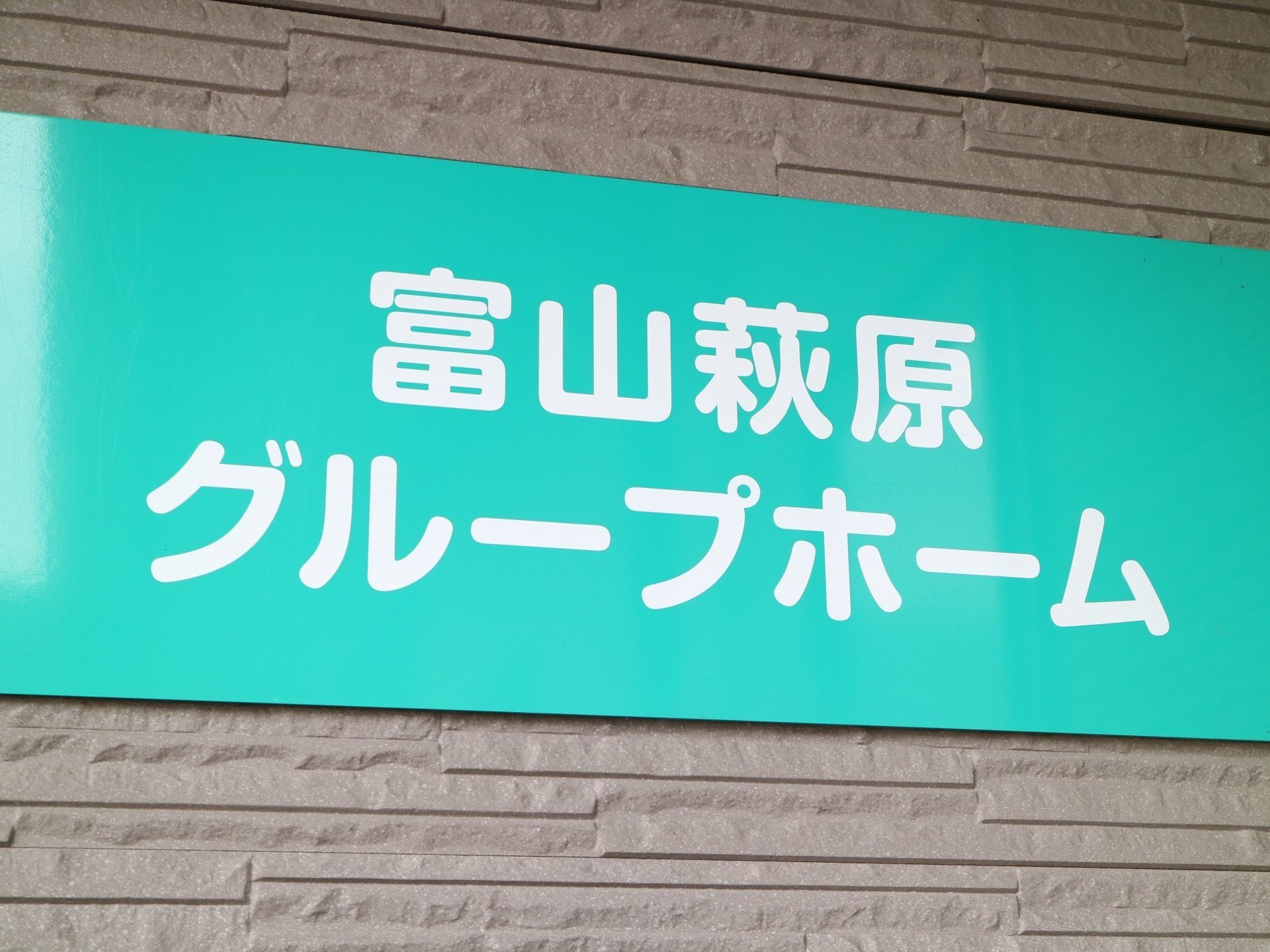グレーの外壁に、大きな看板が取り付けられている。エメラルドグリーンの板に、白い文字で施設名が表示されている。