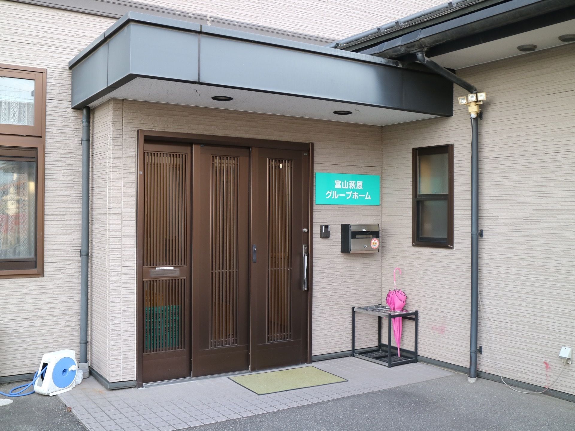 中央に茶色の引き戸が３枚並んでいる。床には黄色い玄関マットが敷かれている。玄関横に傘立てとポストがある。