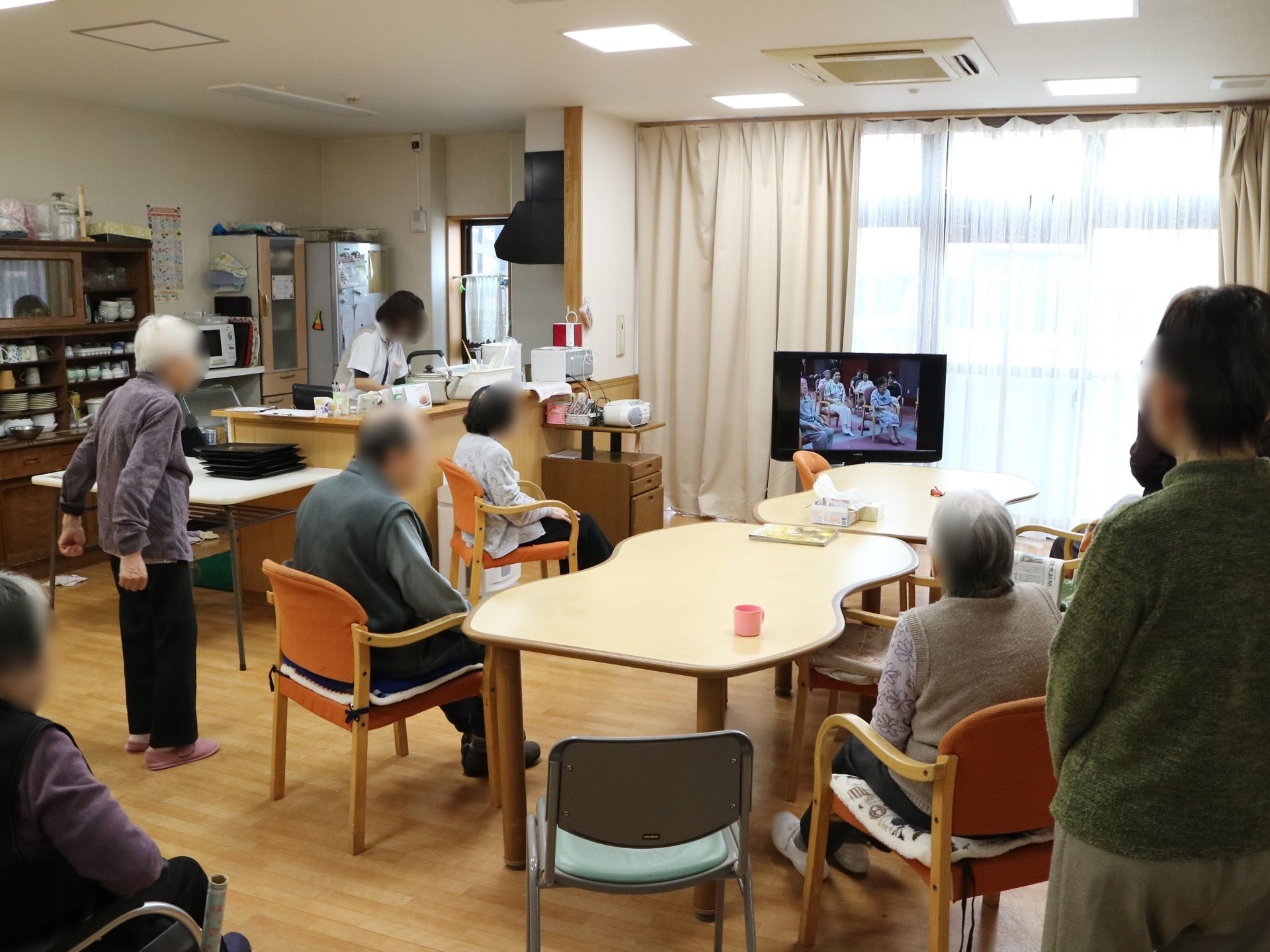 共有スペースには右奥に大きな窓があり、カーテンが掛けられている。左奥にはキッチンがあり、スタッフが作業をしている。