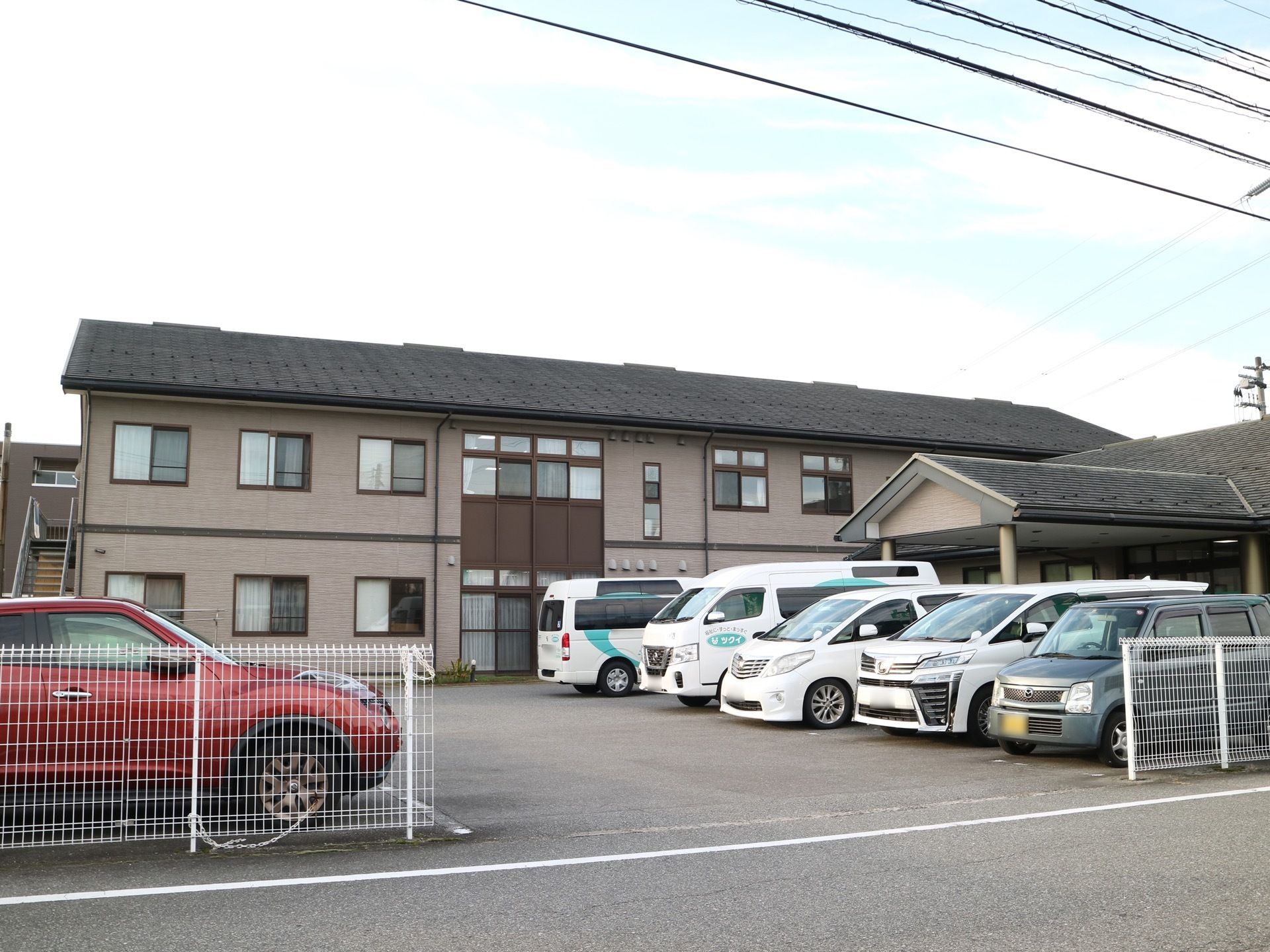 薄い茶色の外壁に黒い屋根の２階建ての施設である。建物の前は駐車場になっており、たくさんの車が停まっている。