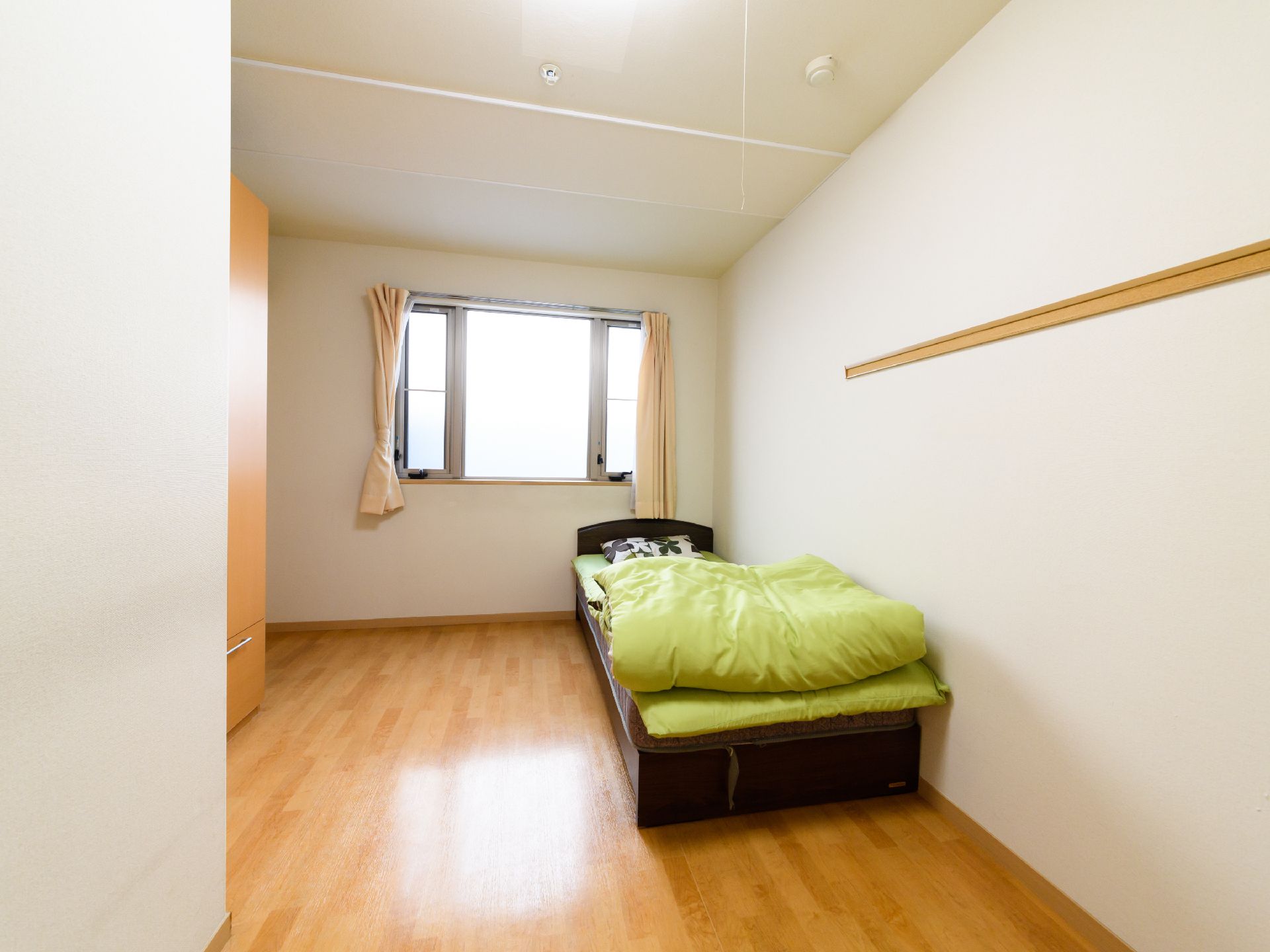 カーテンの付いた大きな窓があるワンルームタイプの明るいお部屋。ベッドを置いてもゆとりのある広さがある。