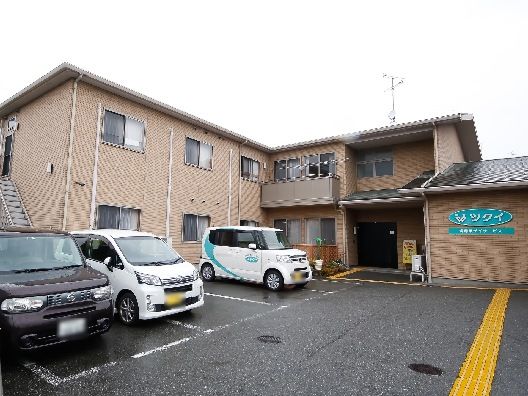 エル字型の２階建ての建物はブラウンの外壁で敷地内には駐車場があり、車が止まっている。