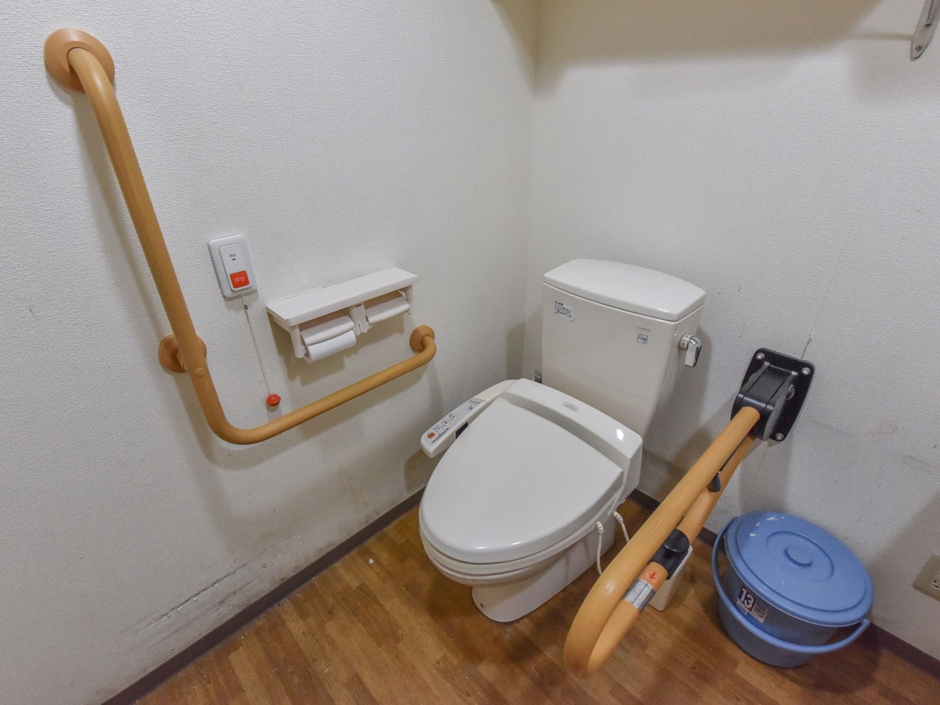 白い壁にフローリング敷の広々としたトイレ。手すりは壁に設置されている他、便器の脇にも可動式の手すりが取り付けられている。