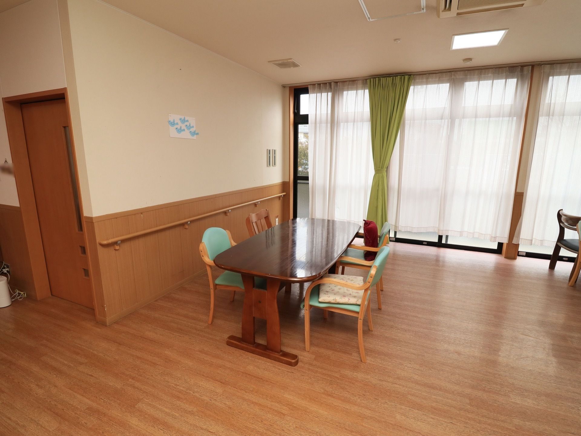 大きな窓から日差しがたっぷり差し込んでいる。椅子とテーブルが置かれている。床はフローリングで余計なものが置かれていない。