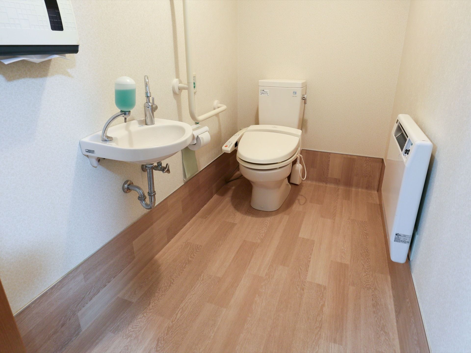 フローリングに白い壁で構成された個室トイレである。中央奥に温水洗浄便座が設置された様式トイレが写っている。