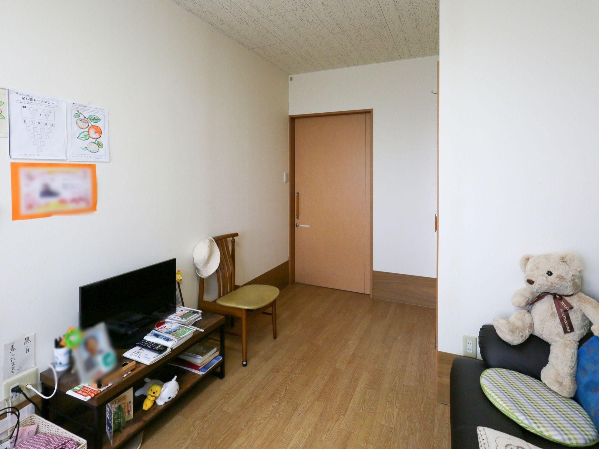 中央奥に廊下へ出る入り口扉が写っている。左手の壁際には大型液晶テレビが置かれた棚と１脚の椅子がある。