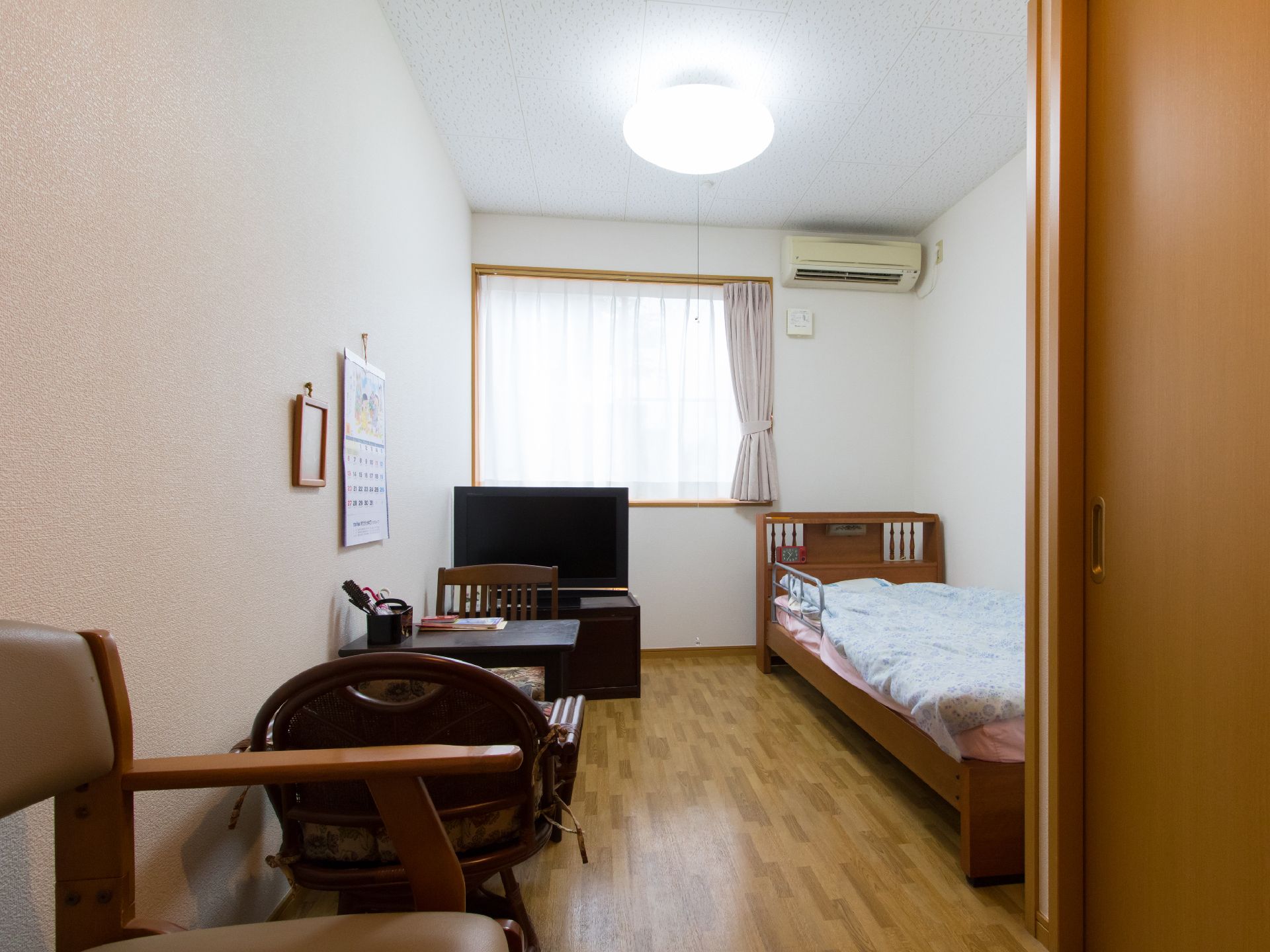 一人用の居室には木目調もベッドがある。テレビがある。腰窓があり陽が差し込んでいる。2人用机が置いてある。