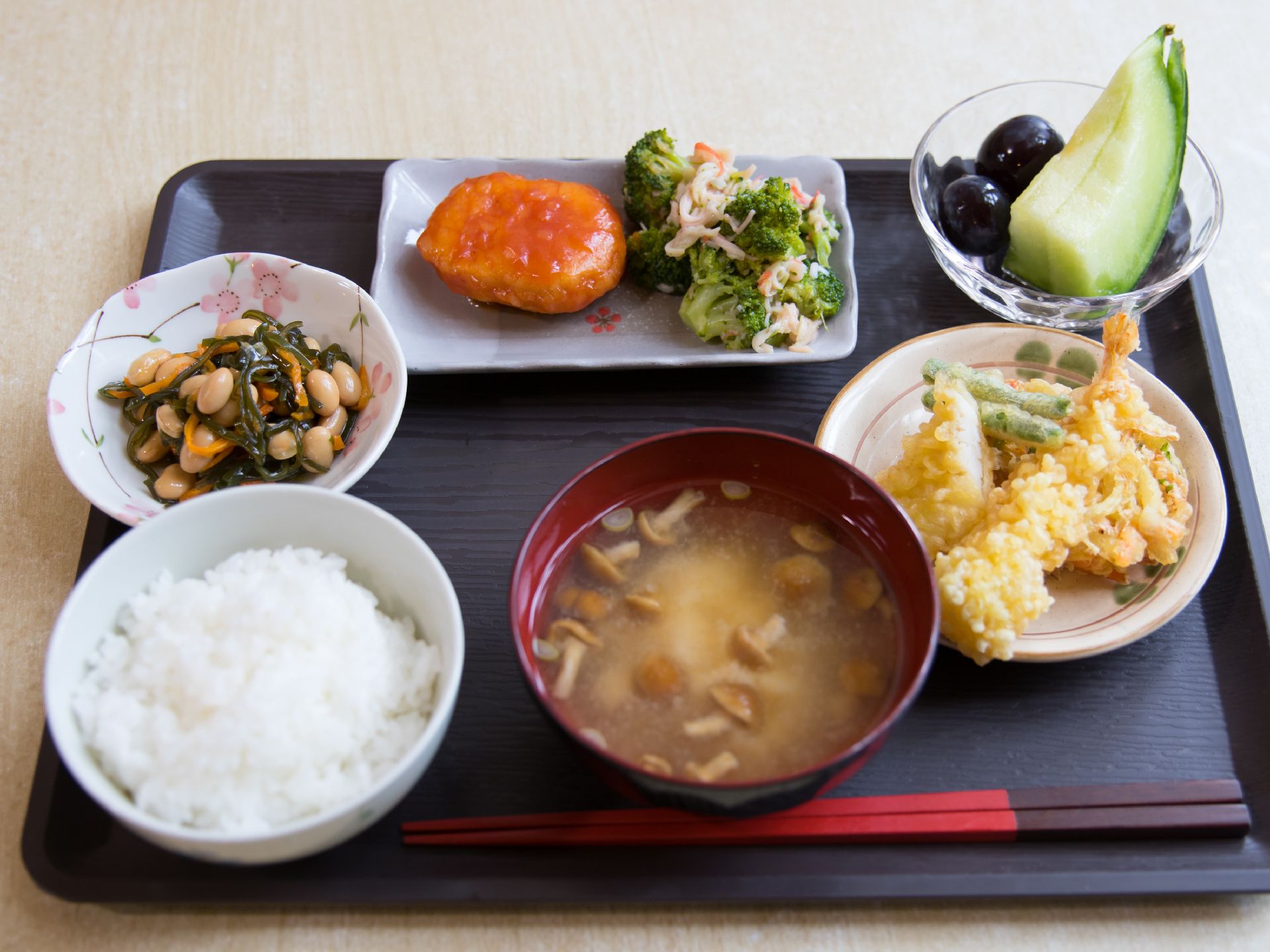 黒いトレーにご飯やみそ汁、天ぷらなど栄養バランスの良い多彩なおかずが並べられている。デザートの果物も置かれている。