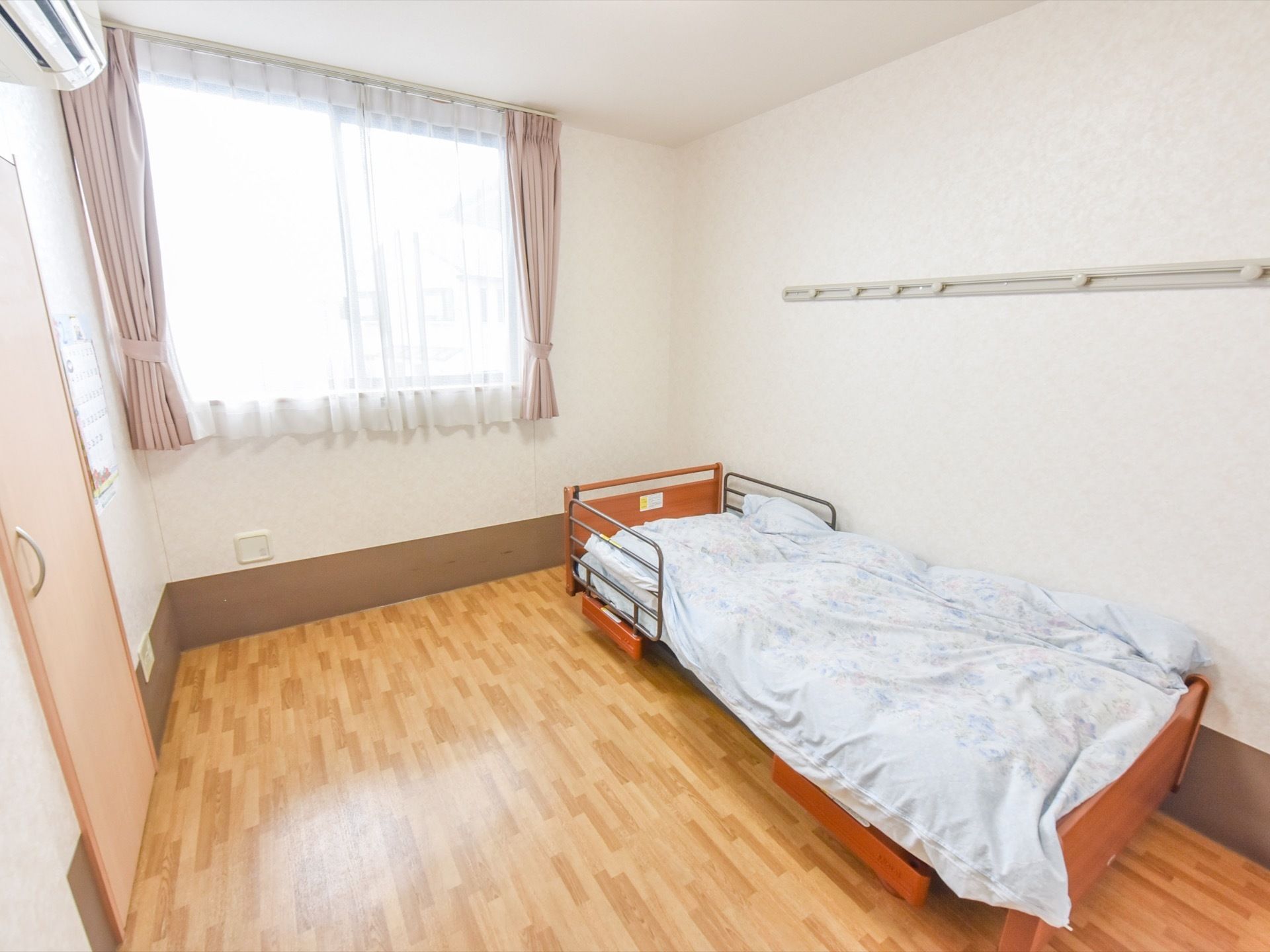 自然光が室内差し込む窓が備わった広い間取りの居室である。エアコンや介護ベッド、レールフックなどが完備されている。