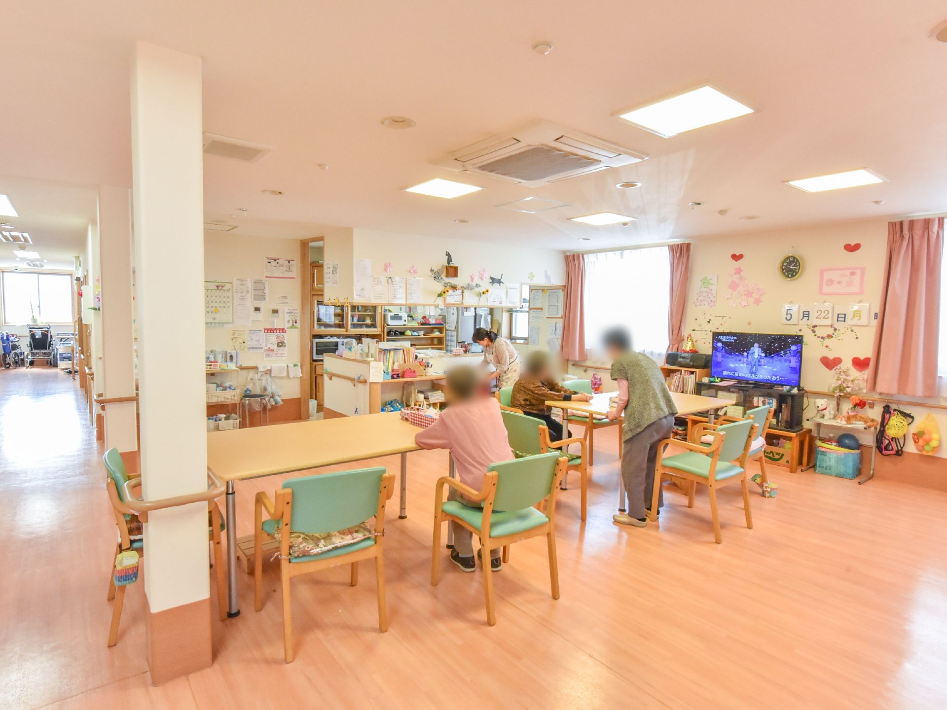 空調のついた広々とした部屋に木目調のテーブルと椅子があり入居者が集まっている。窓からは光が差し込んでいる。