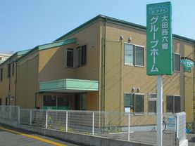 ベージュの壁と緑色の屋根が特徴的な、２階建てのコンパクトな施設である。手前の道路沿いには、施設名称を大きく表示した看板が立てられている。