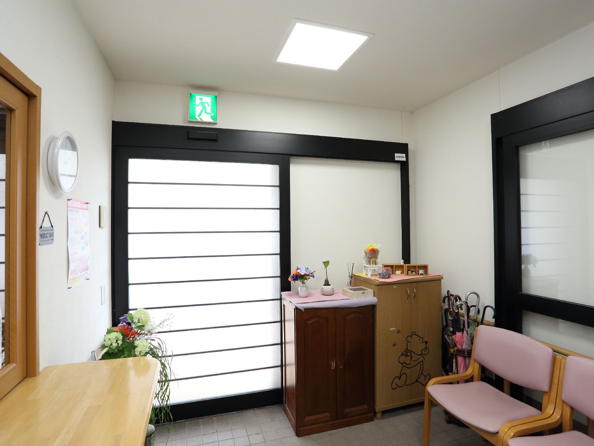 玄関扉は、自動ドアが設置されている。上に小物が飾られた収納家具や傘立て、椅子が2脚置かれている。壁際には、受付が設けられている。