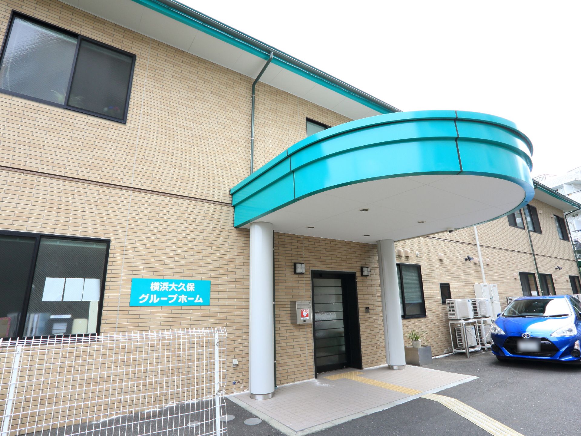 2階建のグループホームは、壁のベージュ色と屋根のブルーグリーンが特徴である。建物前に駐車スペースが設けられている。