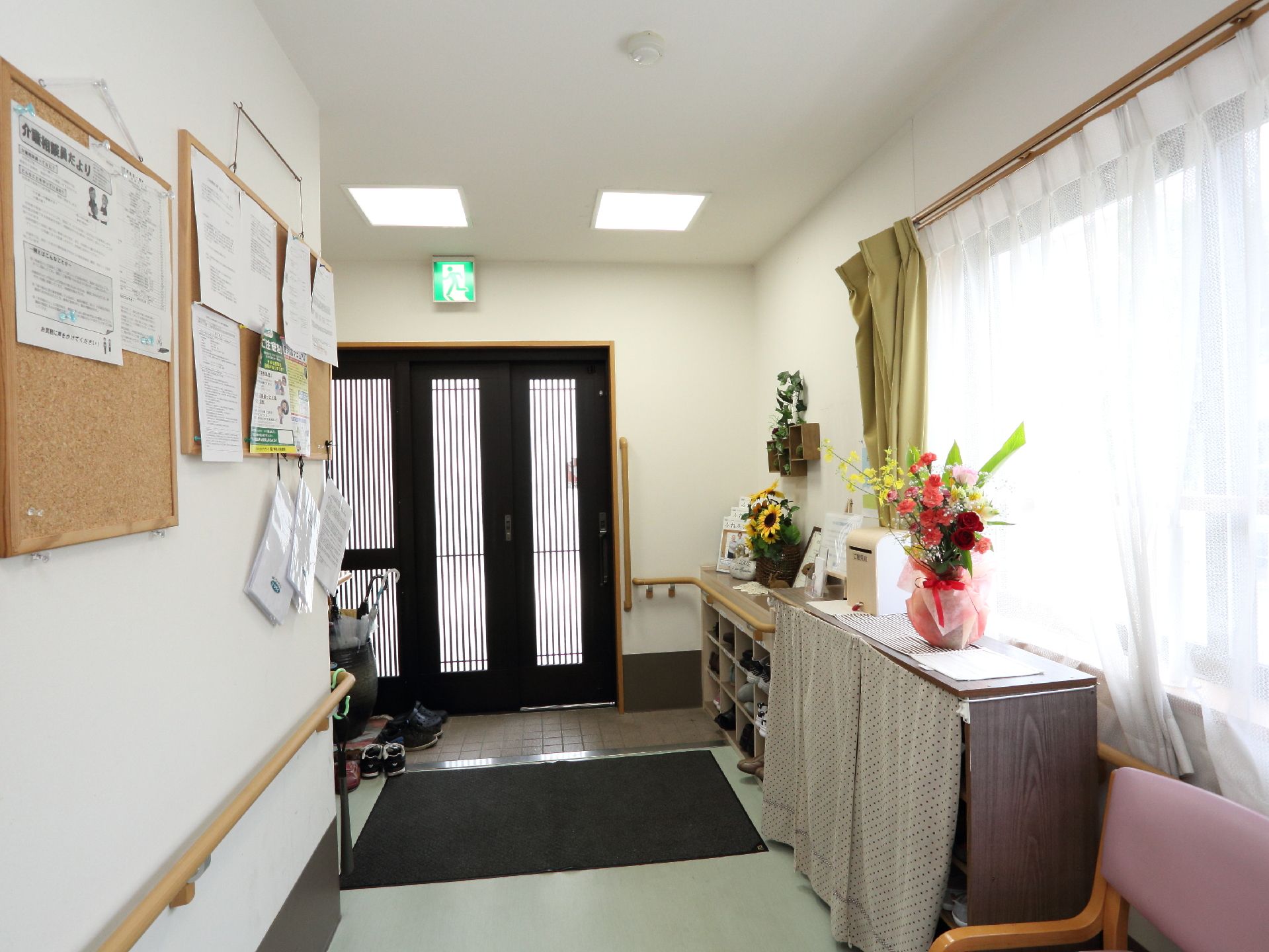 正面に黒い扉のある玄関口の手前には、木製の棚の上に花瓶に入った花が飾られている。奥の靴箱の上にはひまわりの花もある。