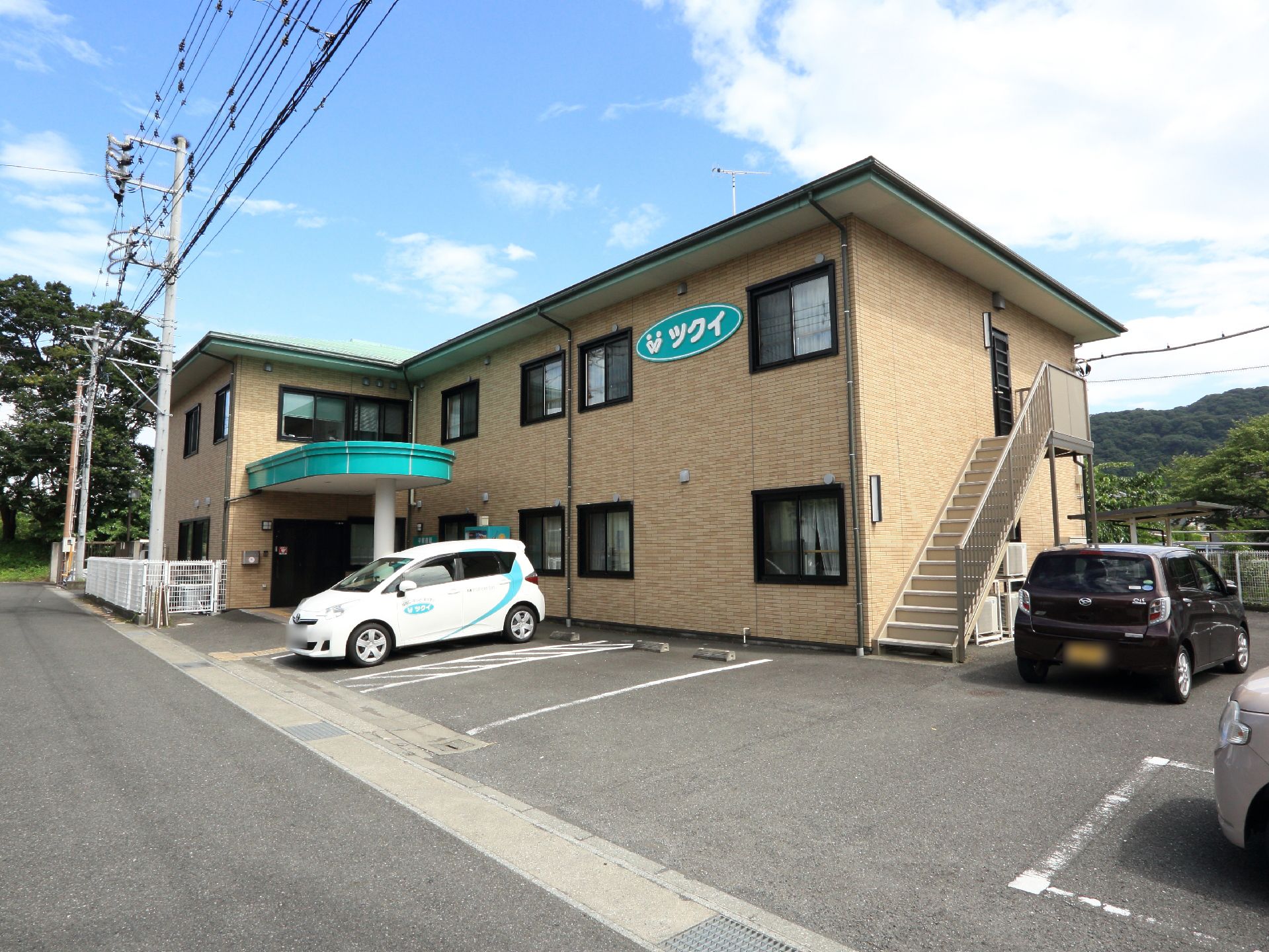 施設の外側部分は、玄関口の上部に緑色の雨除け用屋根がある。そのすぐ側には車が停まっており、こちらは白をベースに緑のラインが入っている。