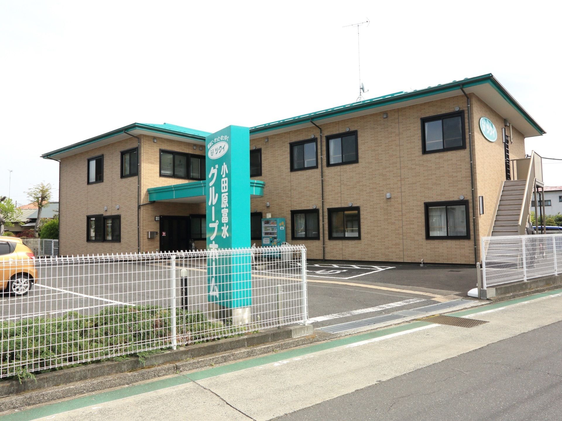 施設の前面には、広く駐車場を完備している。建物の屋根と同色の施設名の入った看板が、道路際のフェンス横に立てられている。