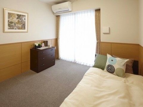 白い壁に木製の腰板が張られている。壁には絵が掛けられており、その斜め下には家具も置いてある。窓の上にはエアコンが付いている。