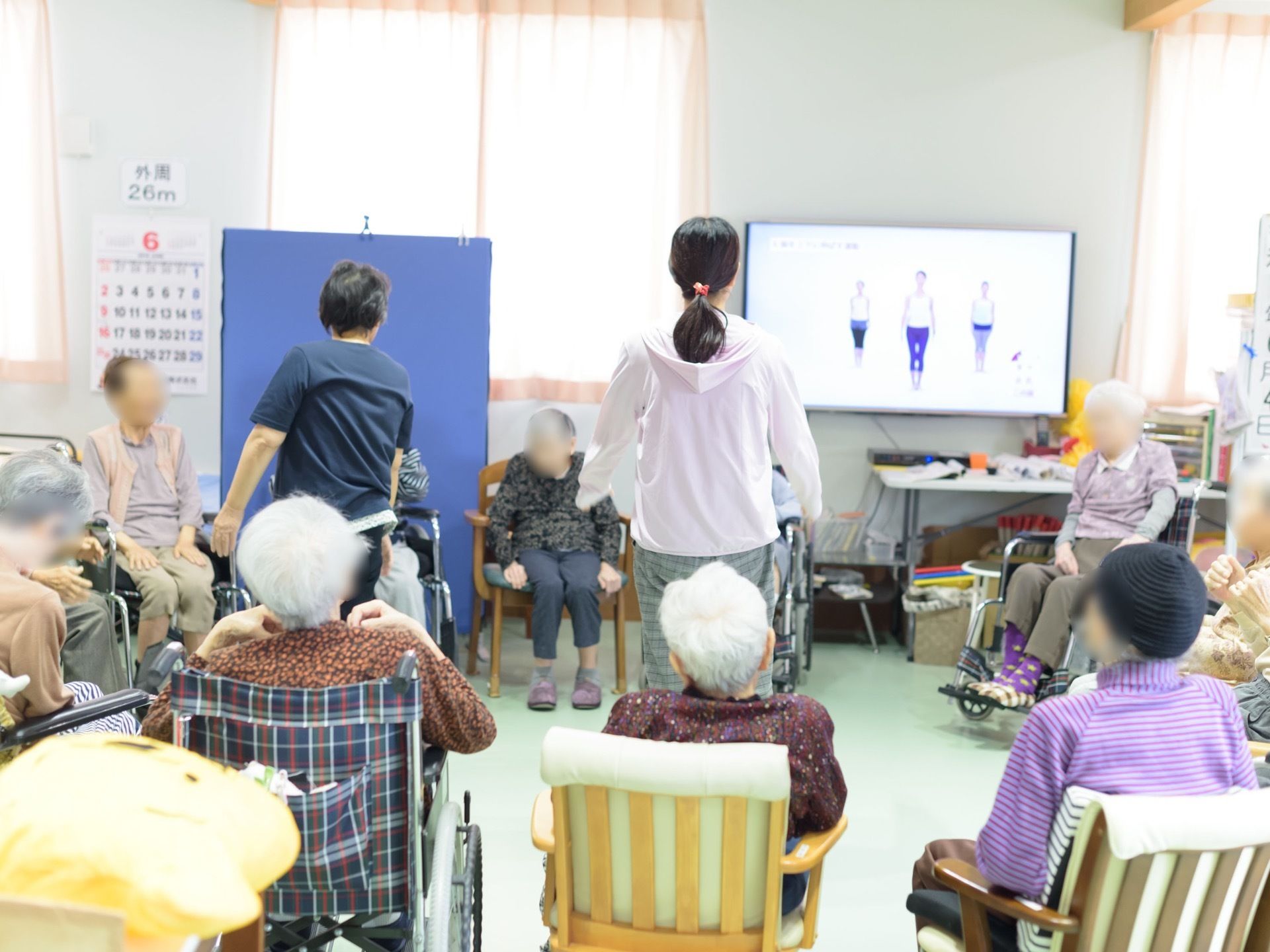 デイサービスの部屋では入居者様が輪になって集まり、テレビ番組やスタッフの先導で座ったまま健康体操を行っている。
