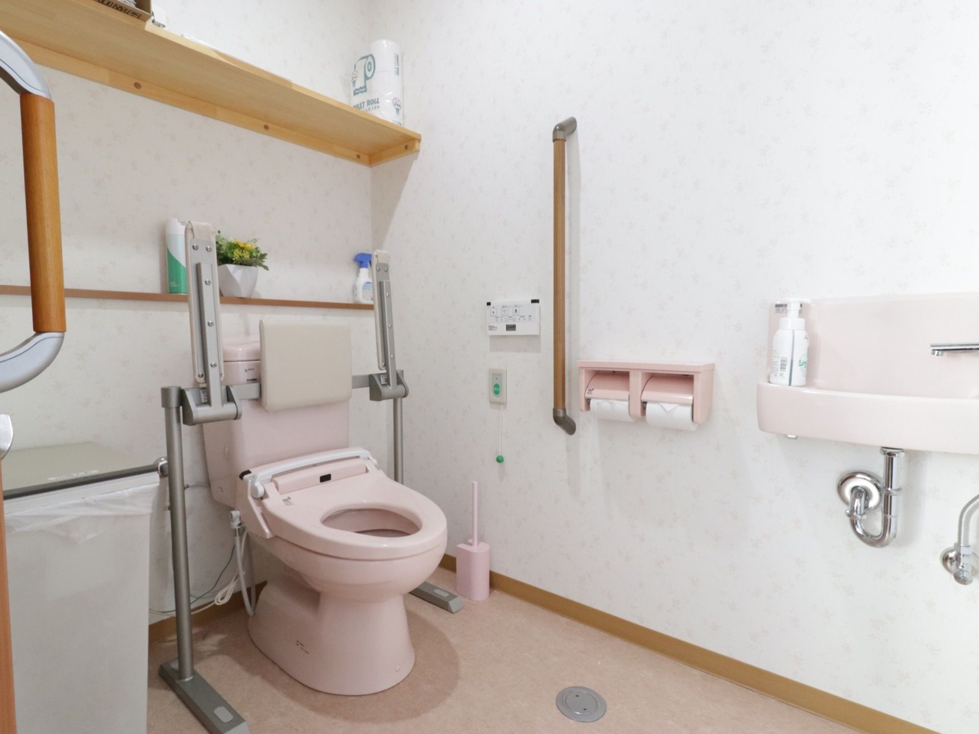 白とピンクを基調とした温かみのあるトイレスペース。コンパクトな洗面台、呼出ボタンが備わっている。便座には手すりと背もたれが付いている。