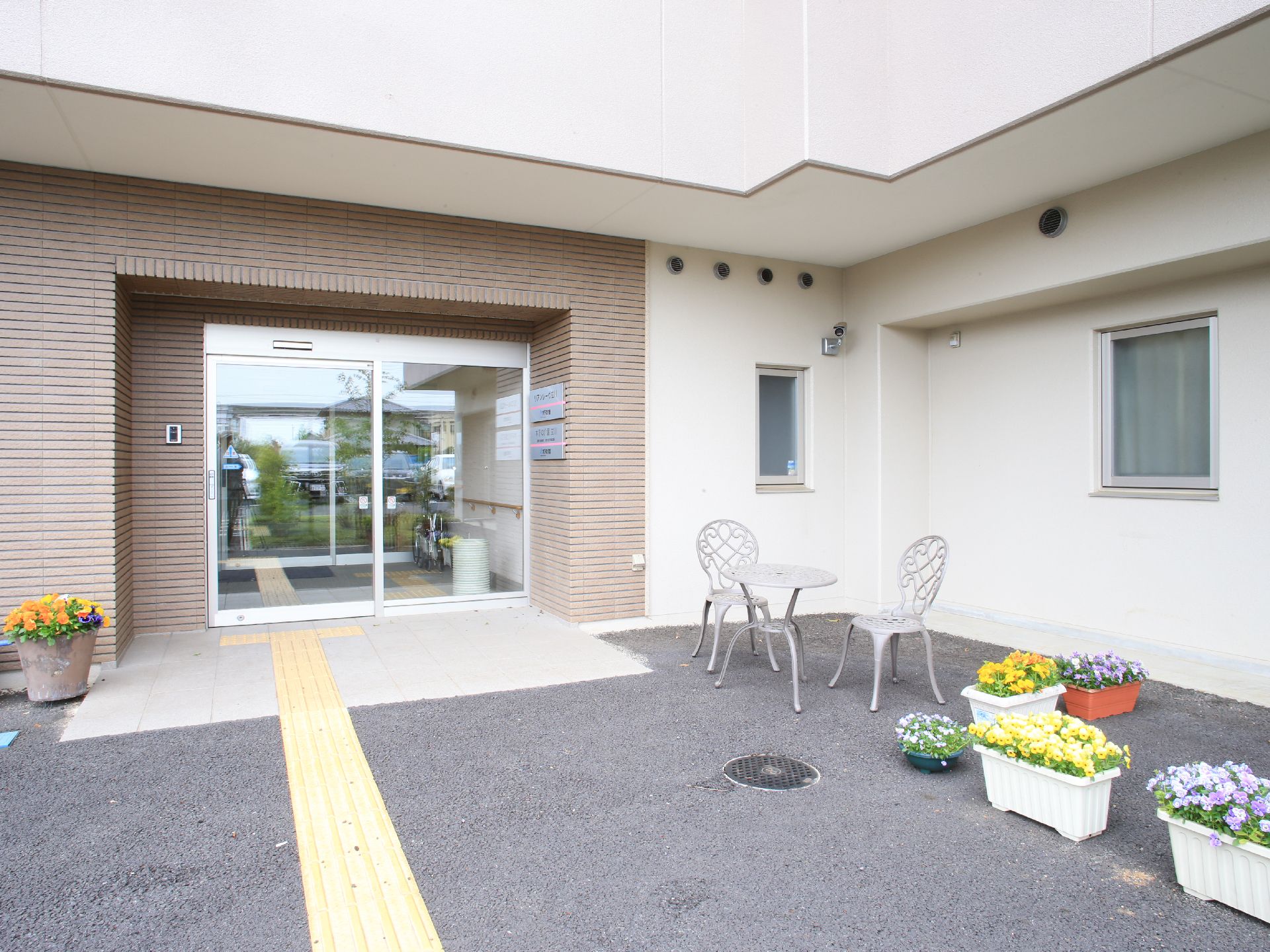 透明な自動ドアの入り口に続くアプローチには、プランターに植えられた色とりどりの花がある。ガーデンテーブルと椅子が置かれている。