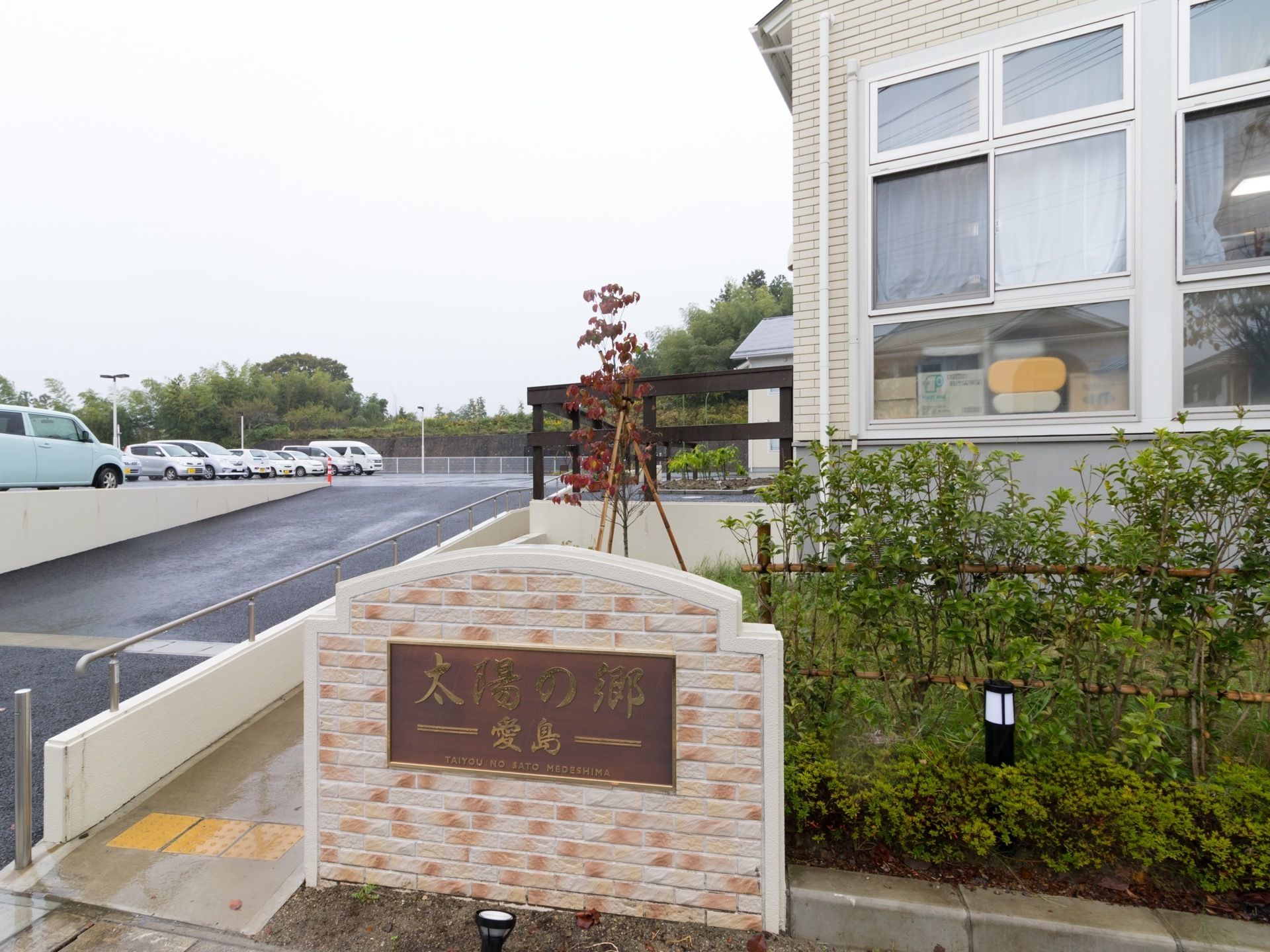 施設名が表示された、レンガ調の看板がある。左手に施設へと続く手すり付き通路があり、右手に緑が植えられている。