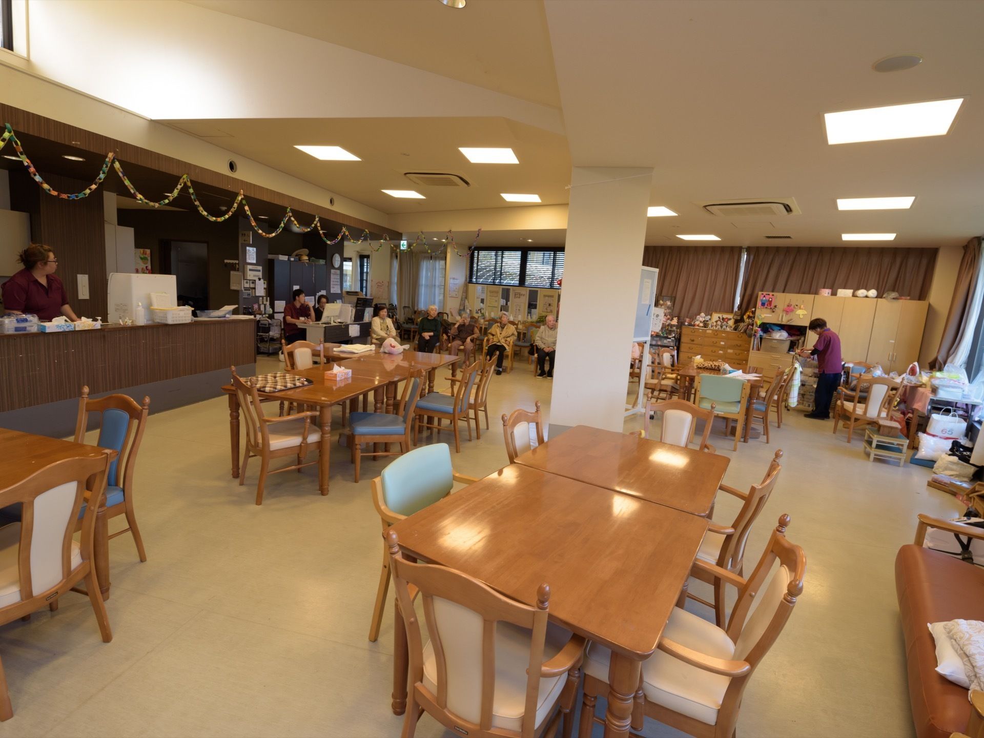 大人数の入居者様が座れるテーブルと椅子が備わっている。壁際には複数の収納棚が置かれ、左側にはキッチンのカウンターが見られる。