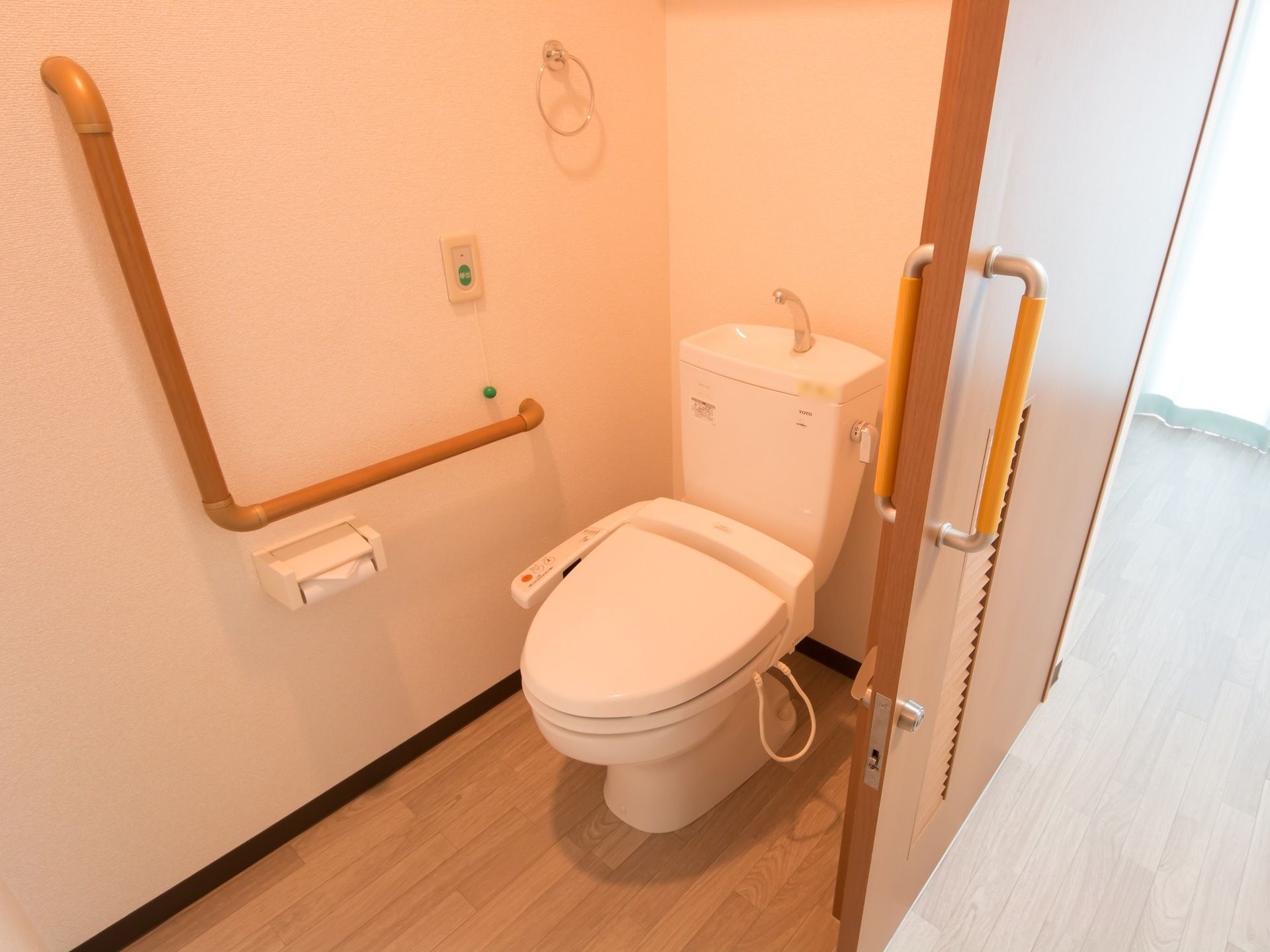 居室内のトイレは、温水洗浄機能付きである。ペーパーホルダーの上には、大型の手すりや、緑色のコールボタンがついている。