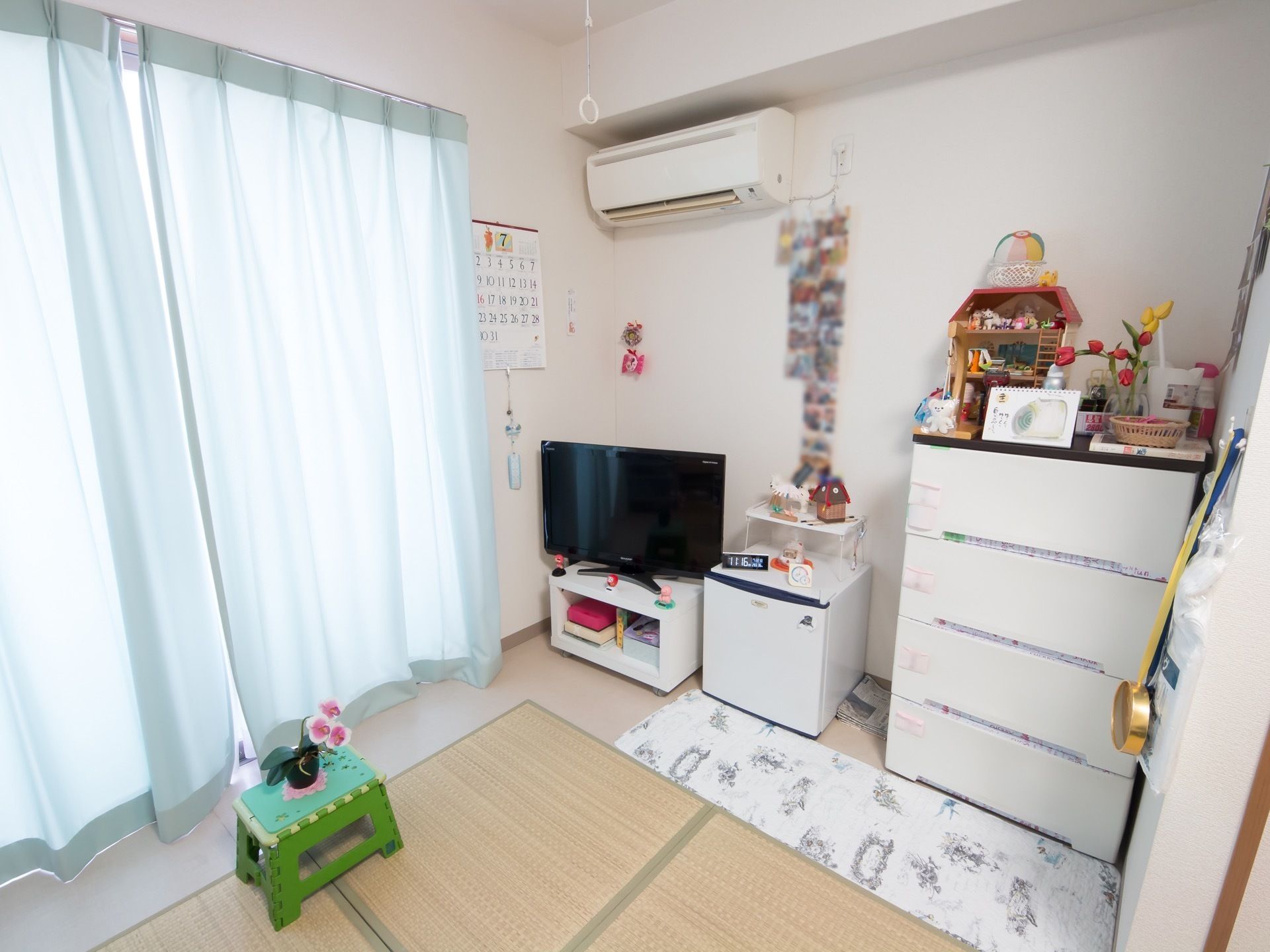 掃き出し窓の右横にカレンダーが掛けてあり、そのすぐ側にエアコンが取りつけられている。黄緑色の台の上に、花が飾ってある。