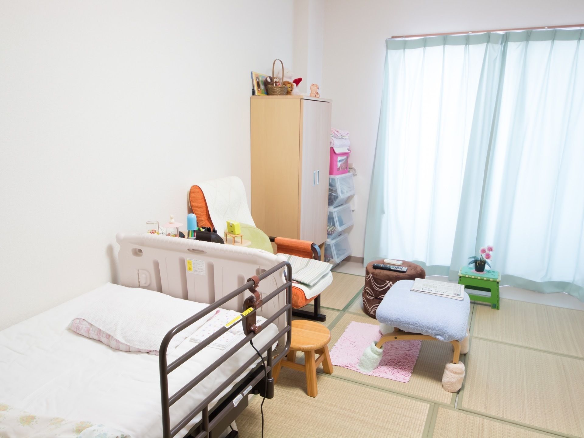 ベージュ色の電動ベッドが壁際に置いてあり、茶色の柵が右端に設置されている。枕の上には、白いカバーが掛けてある。