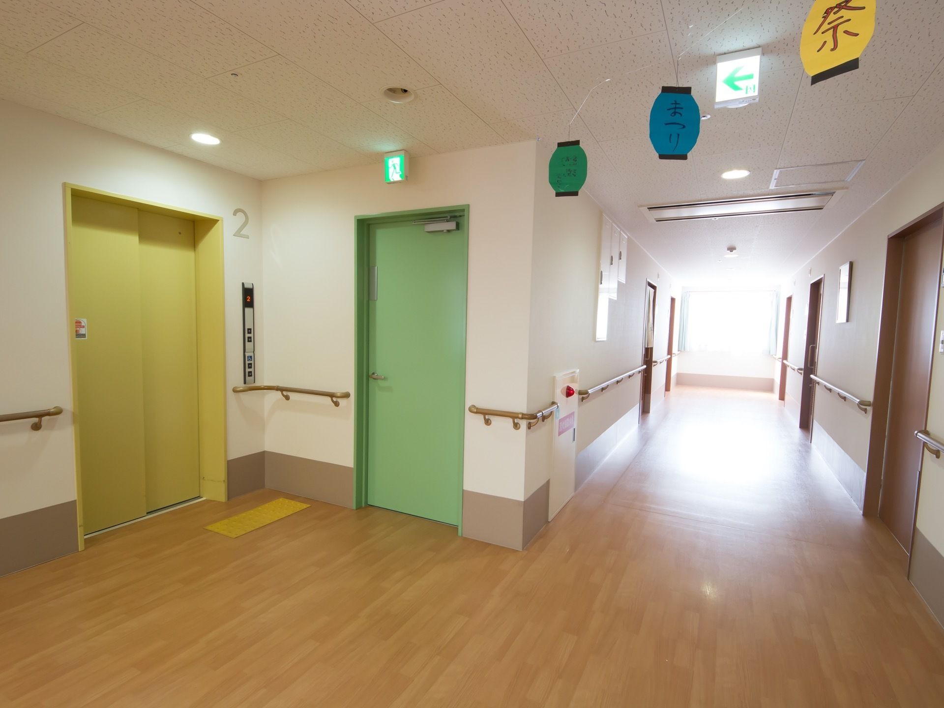 広々としたスペースが確保されたエレベーターホールである。周辺の壁には、ベージュ色の手すりが取りつけられている。