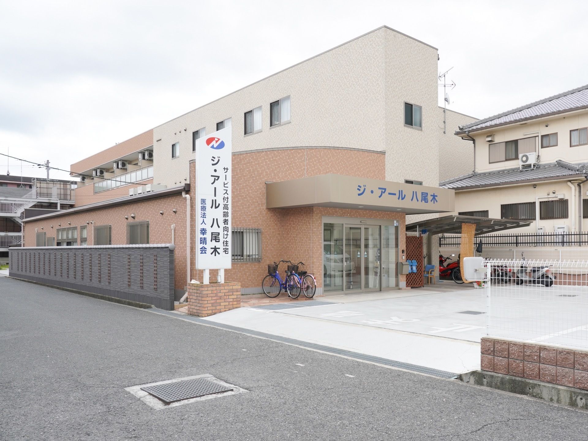 道路と施設はブロック塀やフェンスで区切られている。施設前の路面はアスファルト舗装されており、駐車スペースが確保されている。