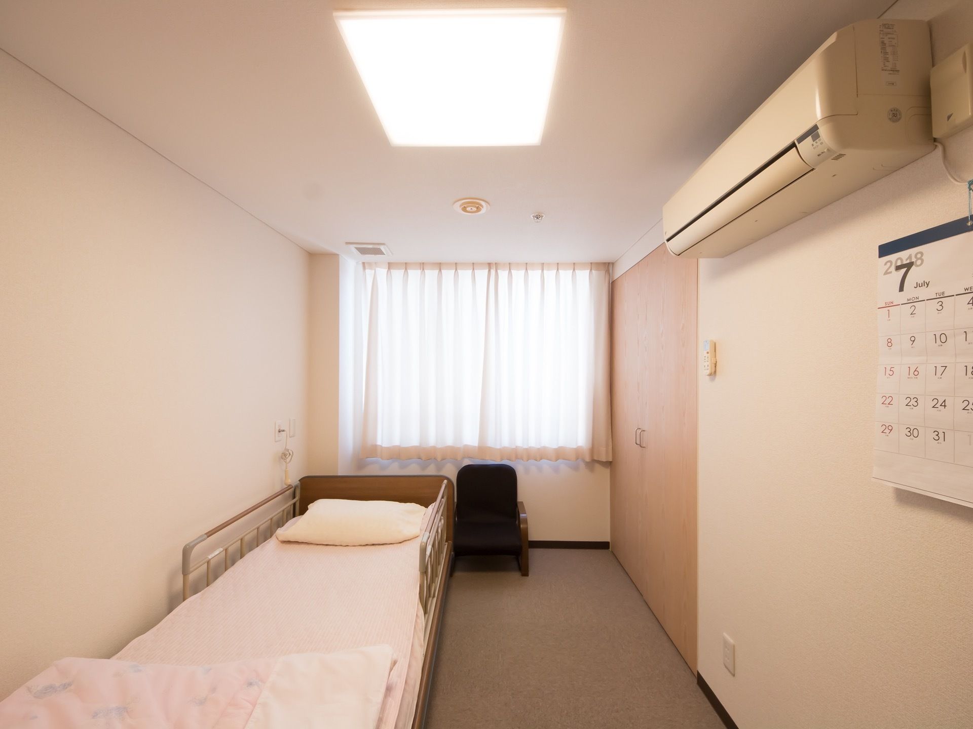 縦長の間取りになっている居室である。ベッドやエアコン、カーテンが完備されている。腰高窓があり、日当たりが良い。