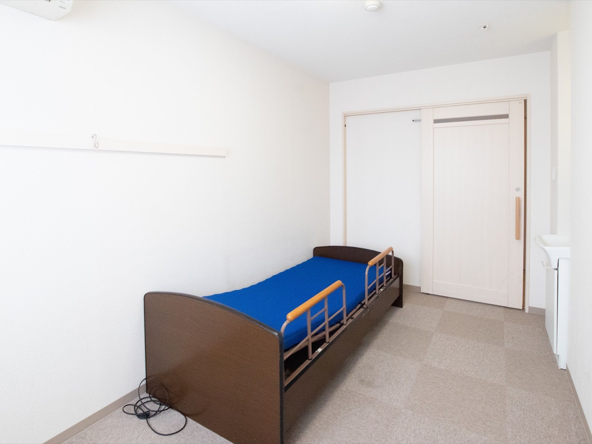 洗面台が完備されている居室である。部屋の壁側には青いマットが敷かれた介護用ベッドが置かれている。床はカーペット素材である。