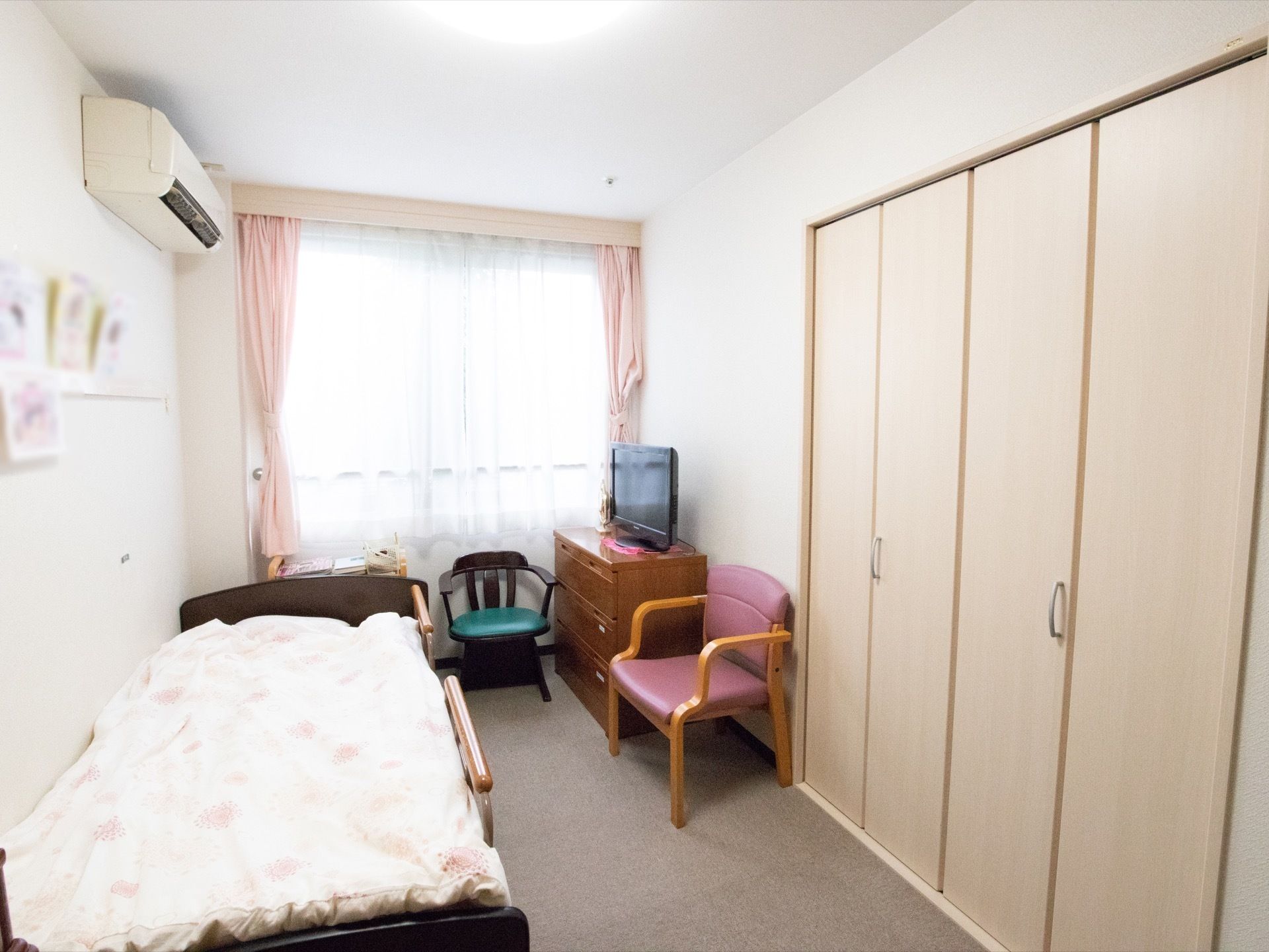 チェストや椅子、介護用ベッドが置かれている居室である。奥側に腰高窓があり、カーテンが掛けられている。壁一面は備えつけの収納になっている。