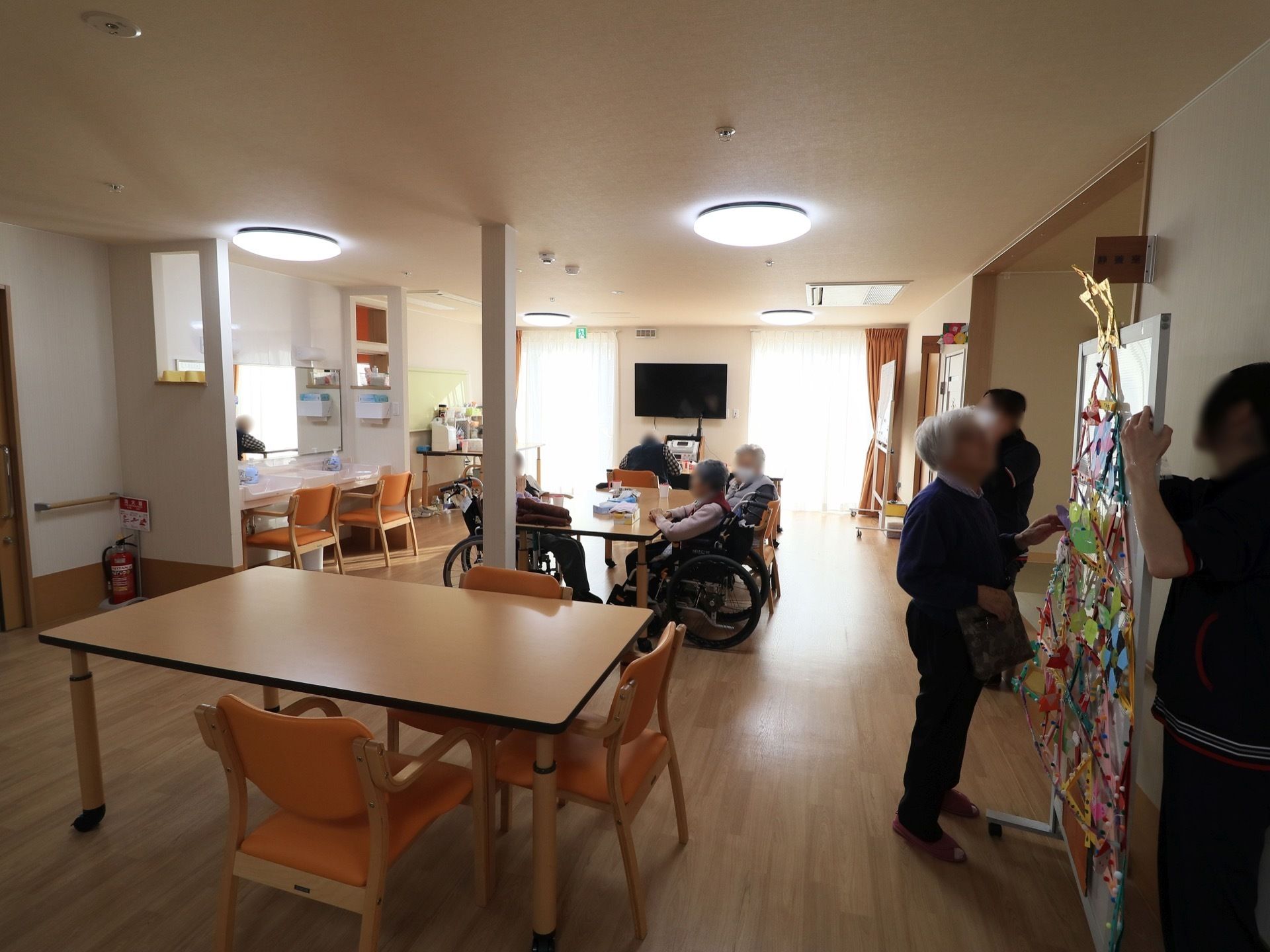 開放的な共有スペースで、入居者様がテーブルに集まり談笑している。ホワイトボードに製作物が飾られている。