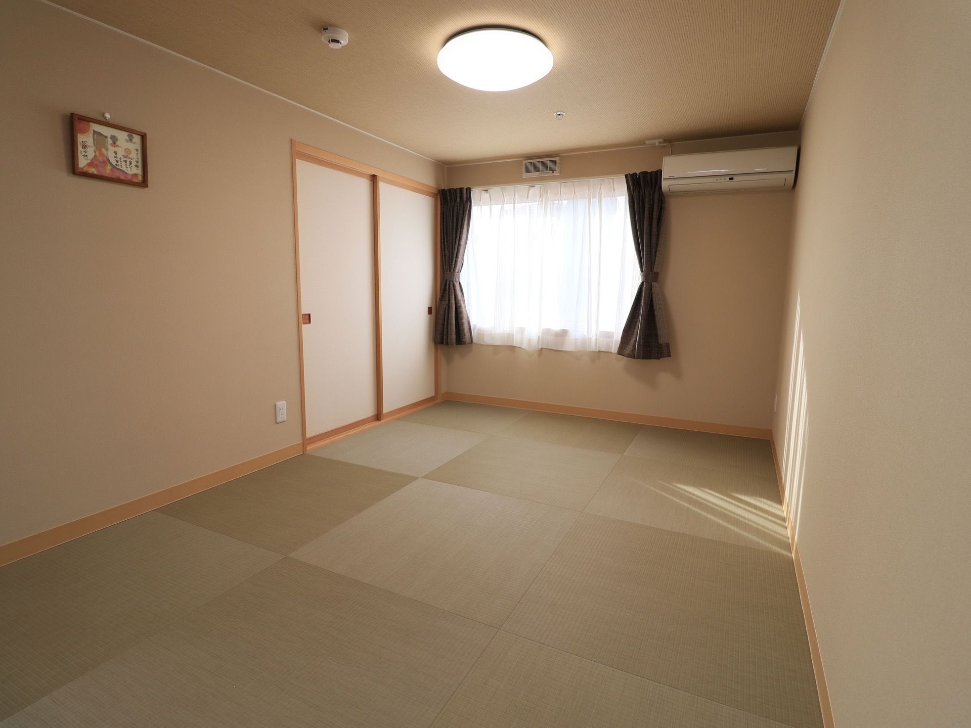 落ち着いた雰囲気の和室である。窓にはカーテンが掛けられている。照明とエアコンが設置されている。壁には絵が飾られている。