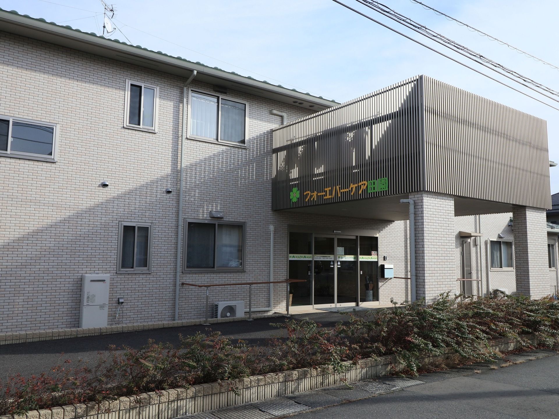 建物はベージュ系の外壁で、落ち着いた雰囲気がある。側には木が植えてあり、スロープの横には花壇がある。