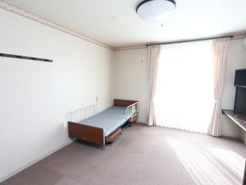 ベッドの他にもテーブルや椅子を置いても十分な空間のある居室である。窓にはピンク色のカーテンがついている。