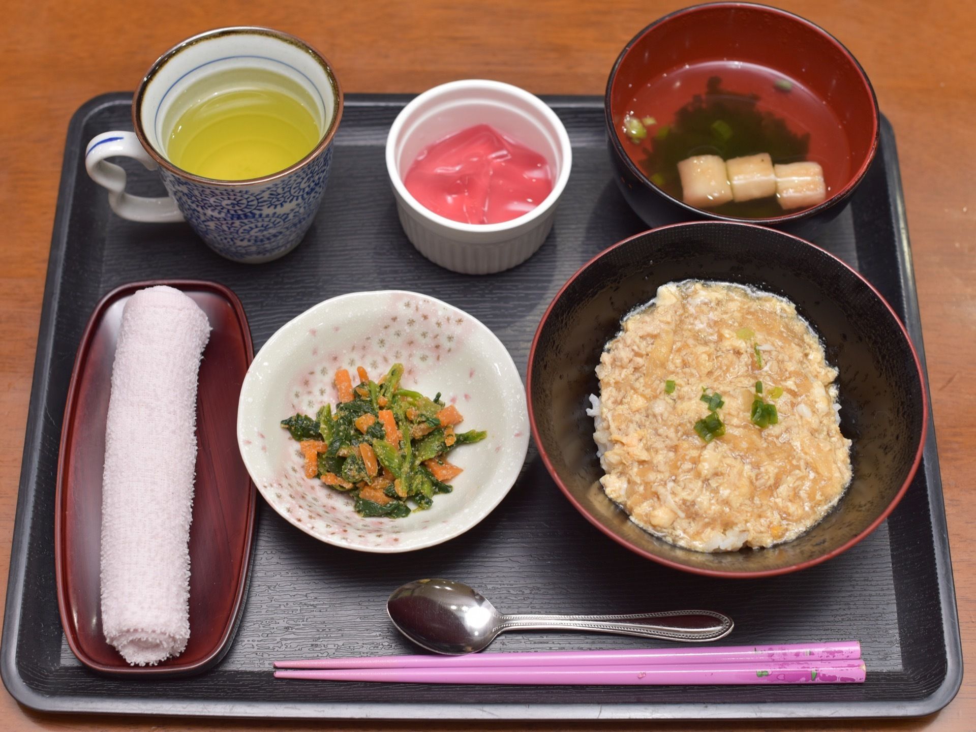 丼物をメインに、小鉢が２種類と汁物が添えられた食事が、黒いトレイに載っている。スプーンと箸が用意されている。