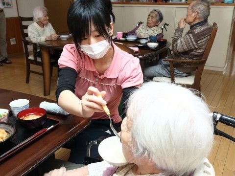 ピンク色の服を着たスタッフに食事を食べさせてもらっている入居者様