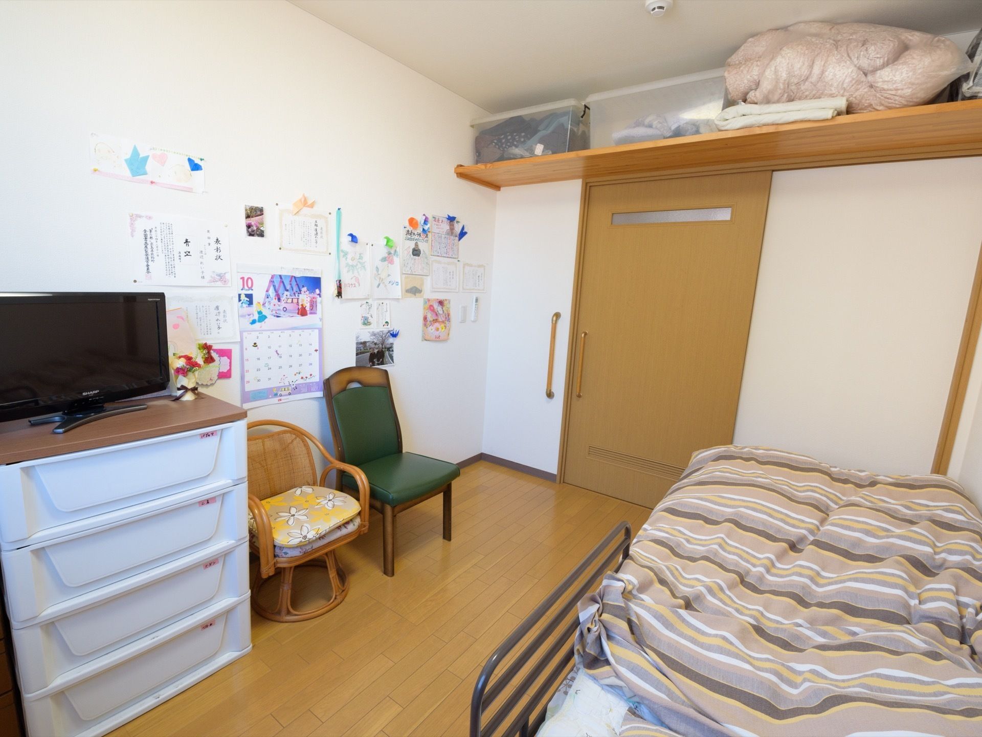 白色の壁に黄土色の出入口ドアと床の居室である。ドアの上部には棚を設けている。壁際にはベッドを置いている。