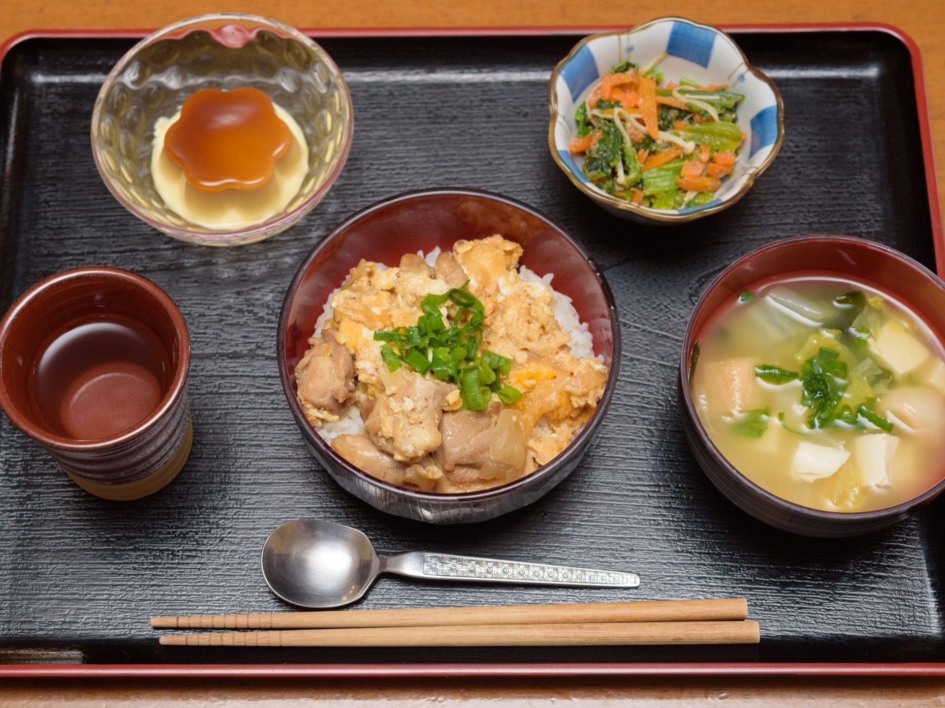鶏肉たっぷりの親子丼と豆腐と野菜、ふのお味噌汁である。野菜の和え物の小鉢とプリンを添えている。手前にスプーンとお箸を置いている。