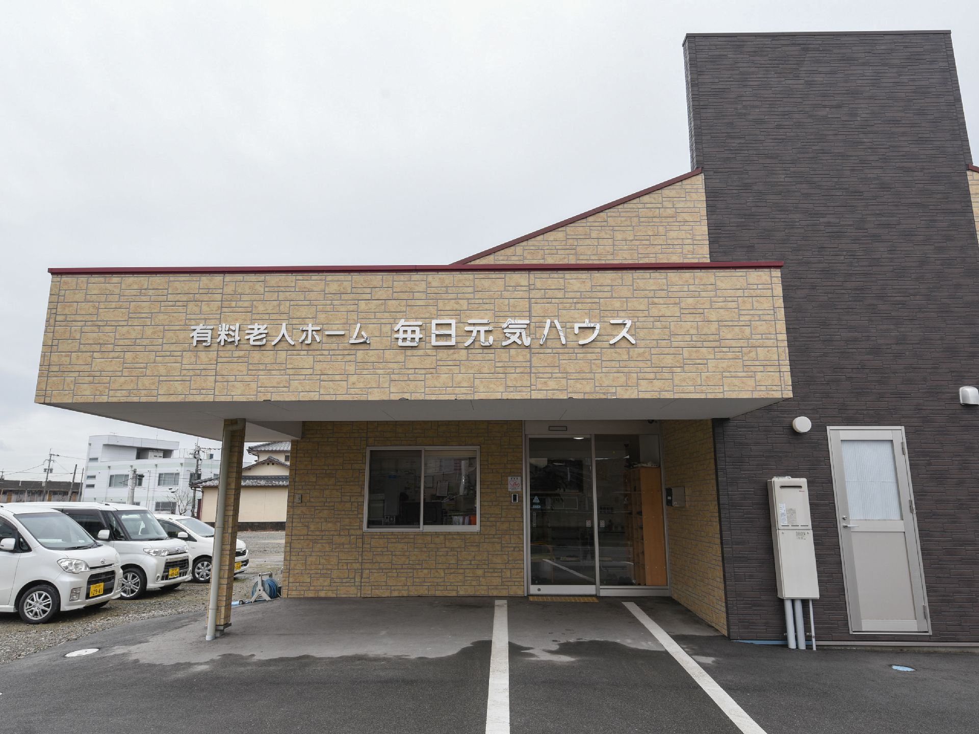 玄関前は大きな屋根が出ている。正面には施設名が大きく表示されている。施設横には駐車場があり軽自動車が停まっている。