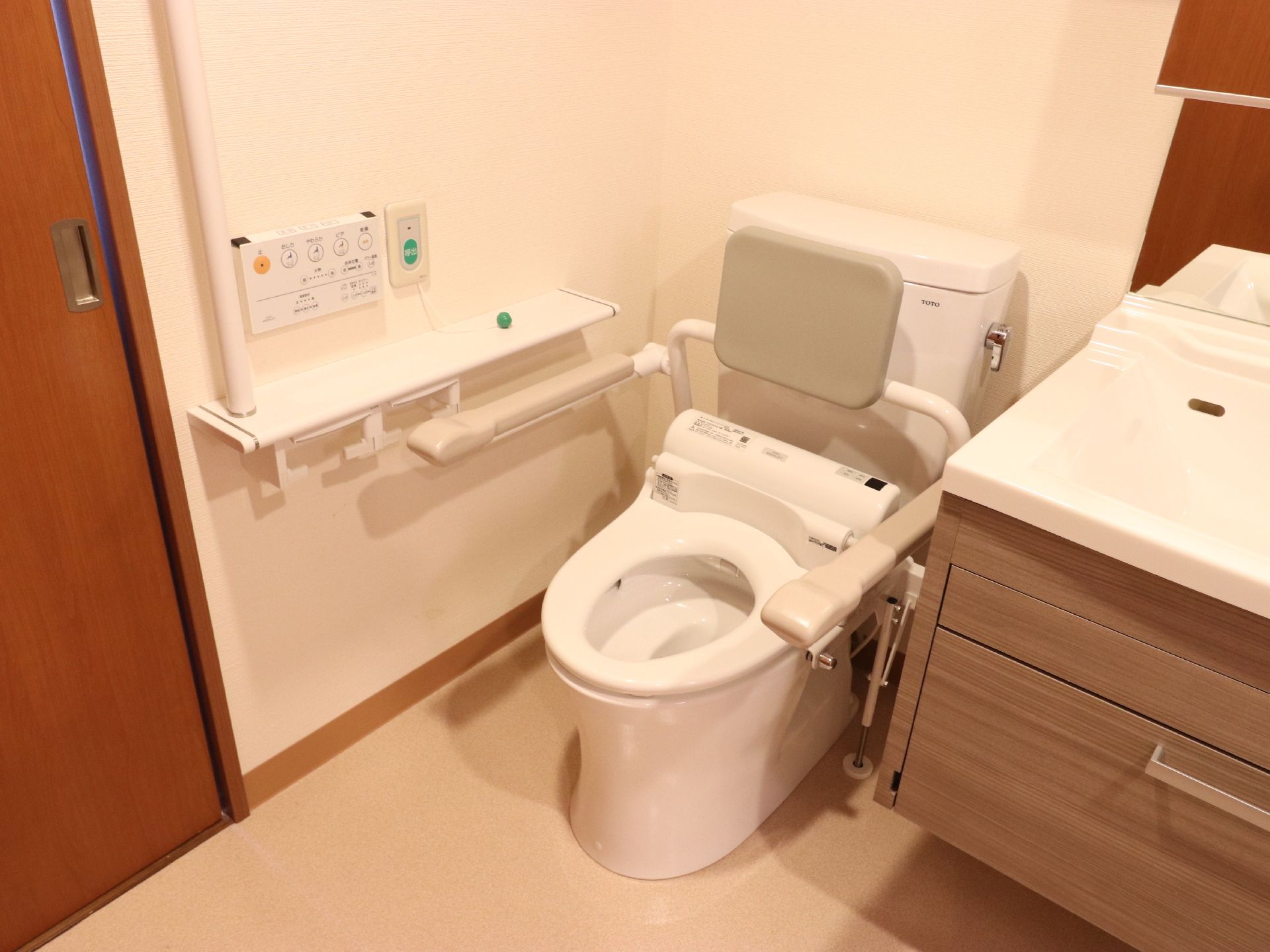 左手のウオッシュレットトイレには背もたれと手すりがついている。操作パネルとナースコールが壁に取り付けられて今sう。