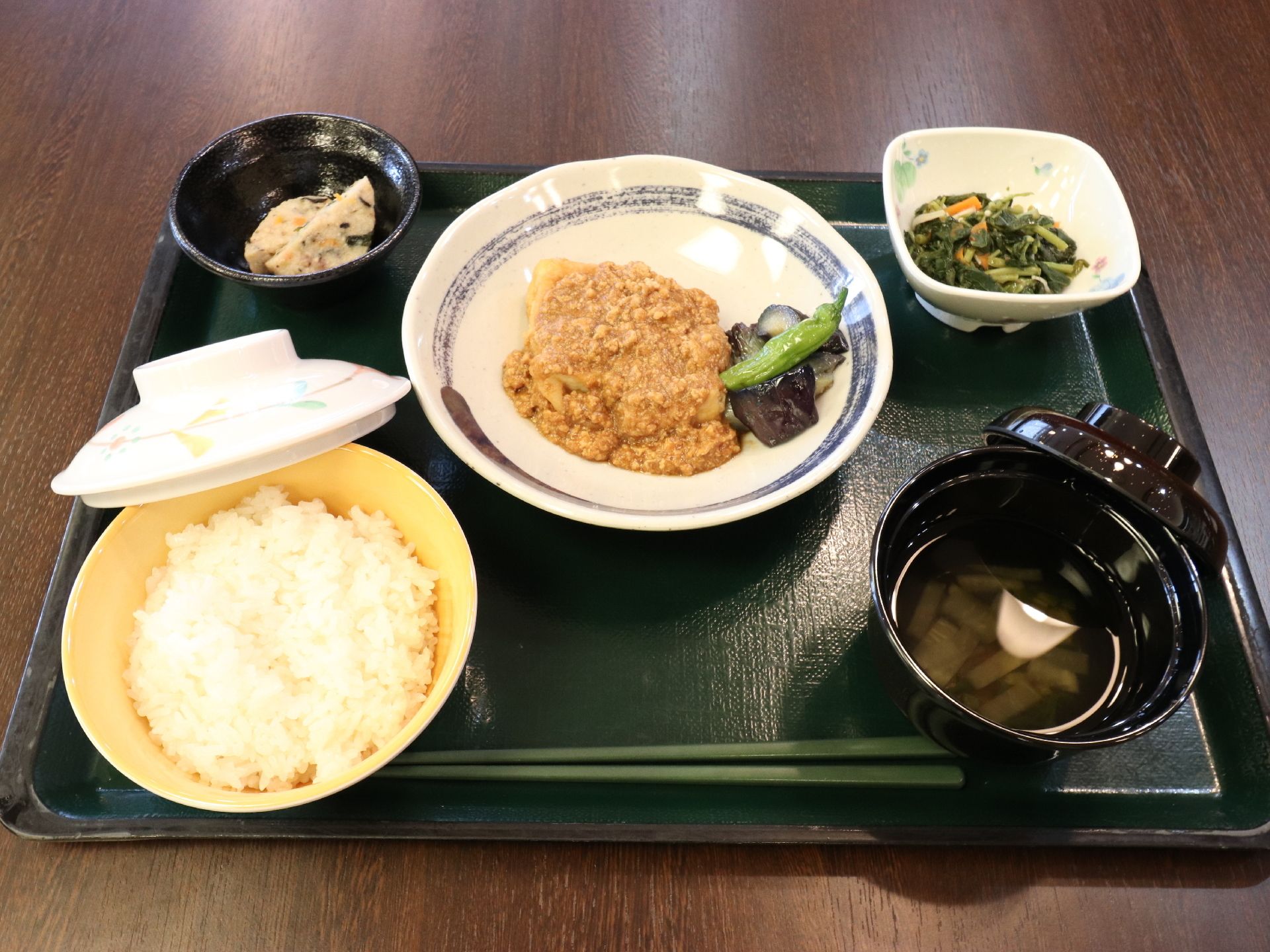 黒いトレイに食事が用意されている。野菜に肉そぼろあんかけがかかった主菜に副菜が2点、お吸い物にご飯が提供されている。