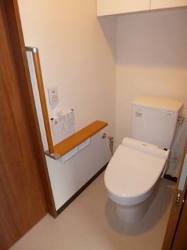 洋式便器が設置されており、便器の上には収納棚がある。扉横には縦型の手すり、ペーパーフォルダー上には棚がある。