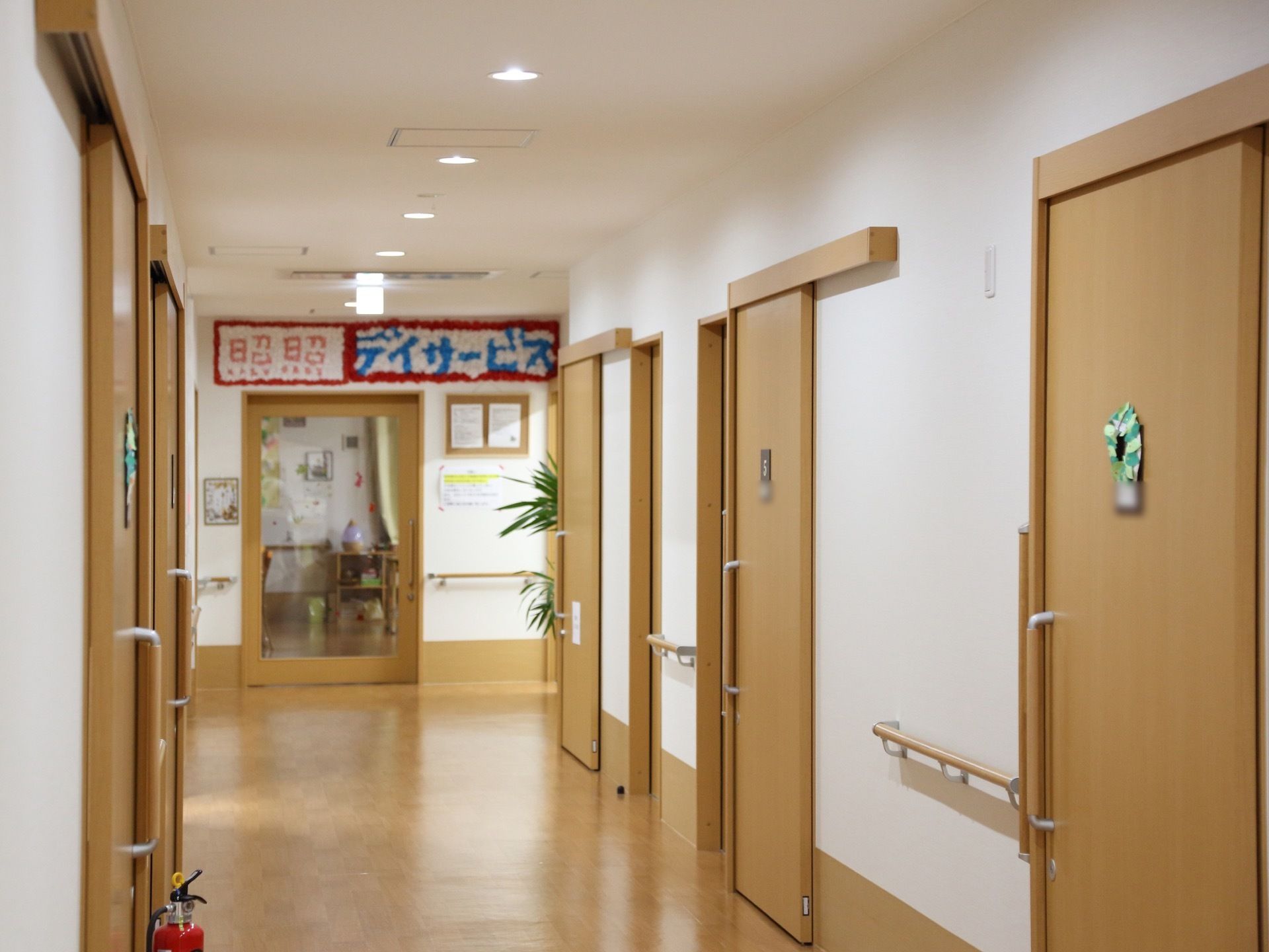 通路の奥にはデイサービス室があり、色とりどりのペーパーフラワーで飾り付けされている。出入口のドアは、ガラス張りになっている。