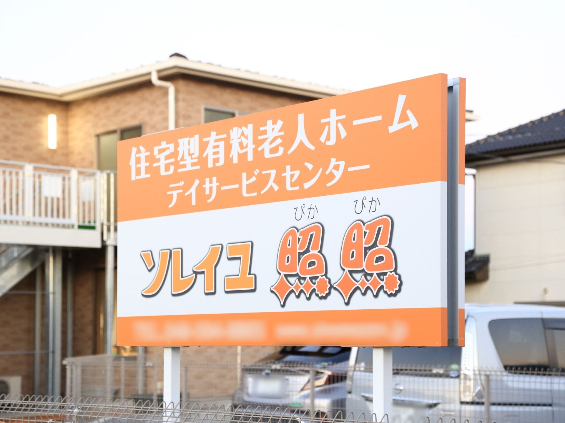 施設の建物の斜め前あたりに、大型の看板が取り付けられている。オレンジ色と白のポップな色合いである。後方には車が置いてある。